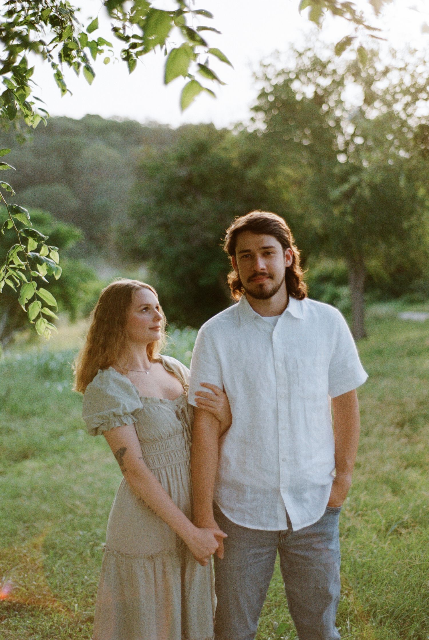 A young couple stands in a lush green field at golden hour, the woman in a sage dress and man in a white linen shirt.