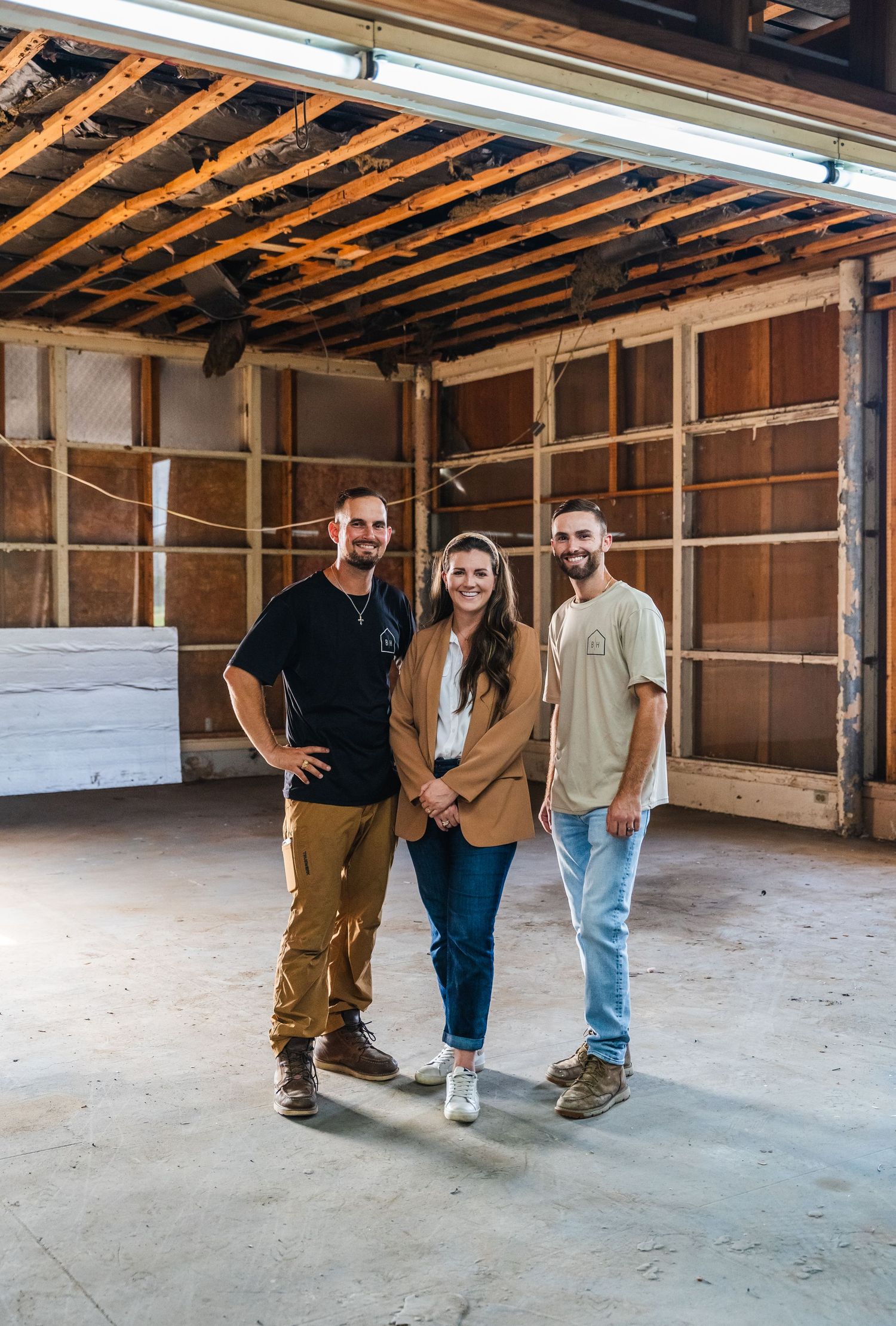 Three construction professionals smiling in a gutted renovation space with exposed wood framing and concrete floors.