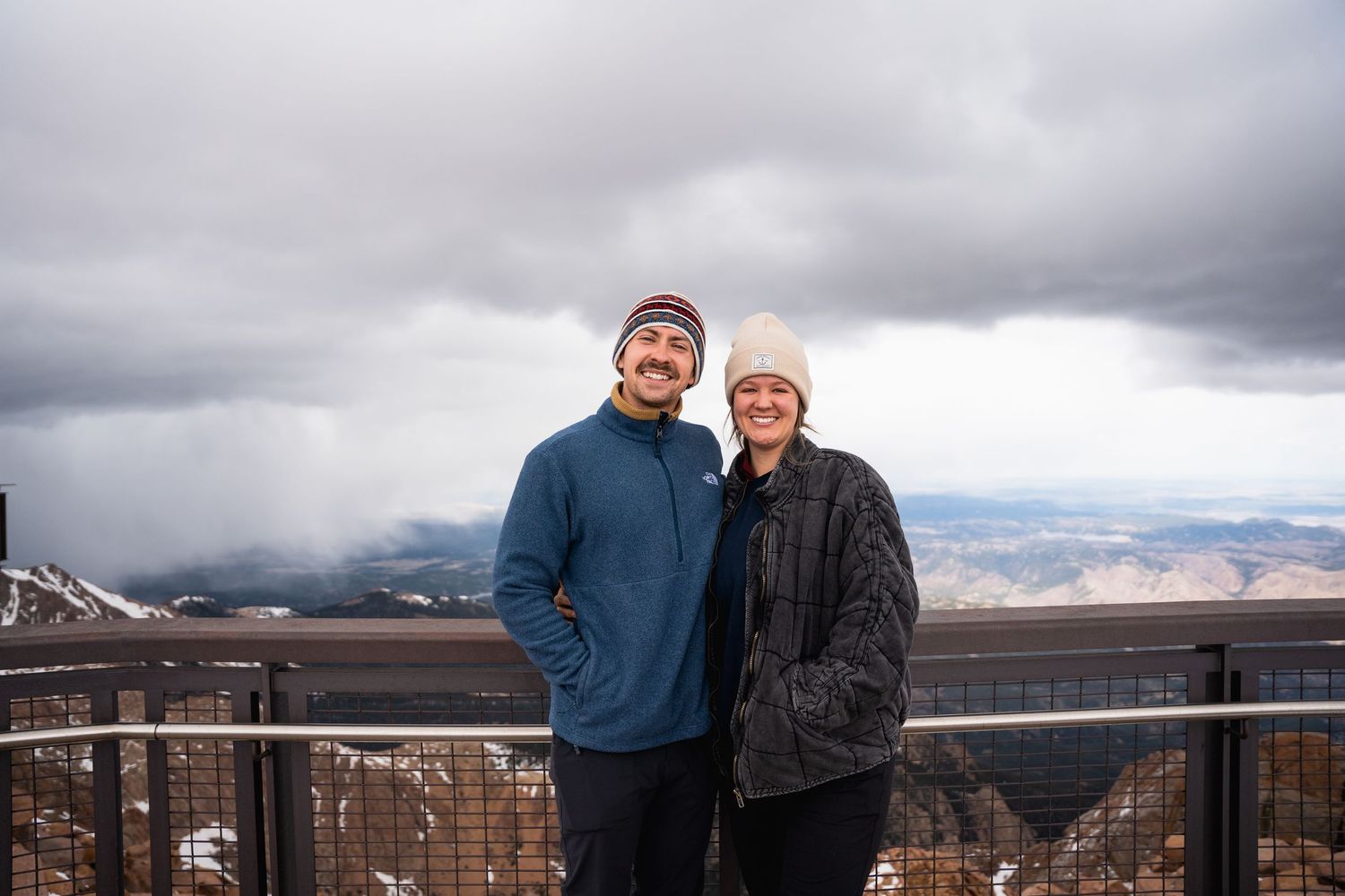 Smiling couple in winter hats pose at a mountain overlook with dramatic cloudy skies behind them.