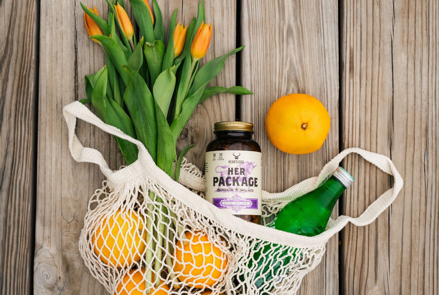 Mesh bag with oranges, tulips, a green bottle, and Her Package supplement on a wooden surface.