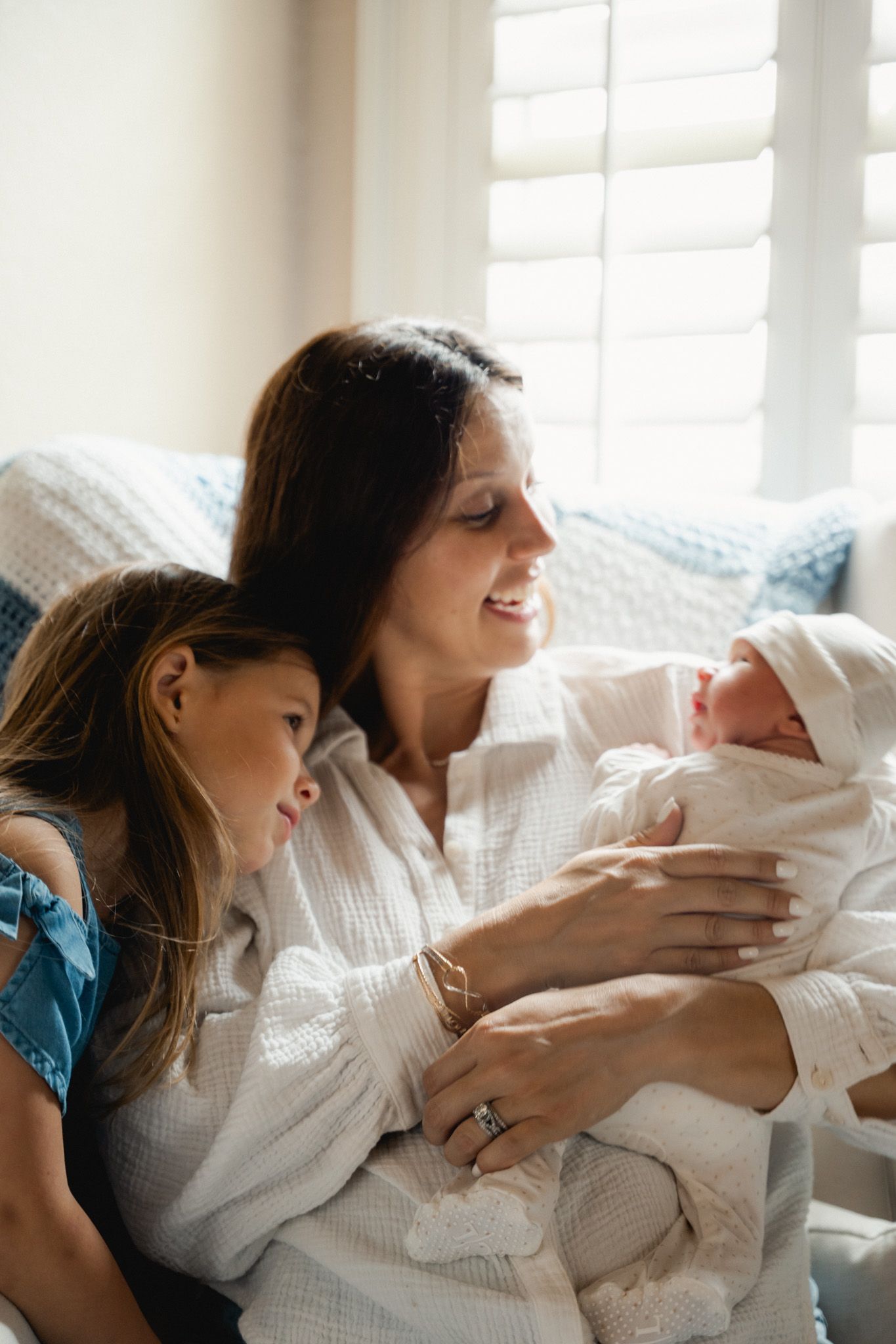 Smiling mother holding newborn baby with older daughter looking on by window.