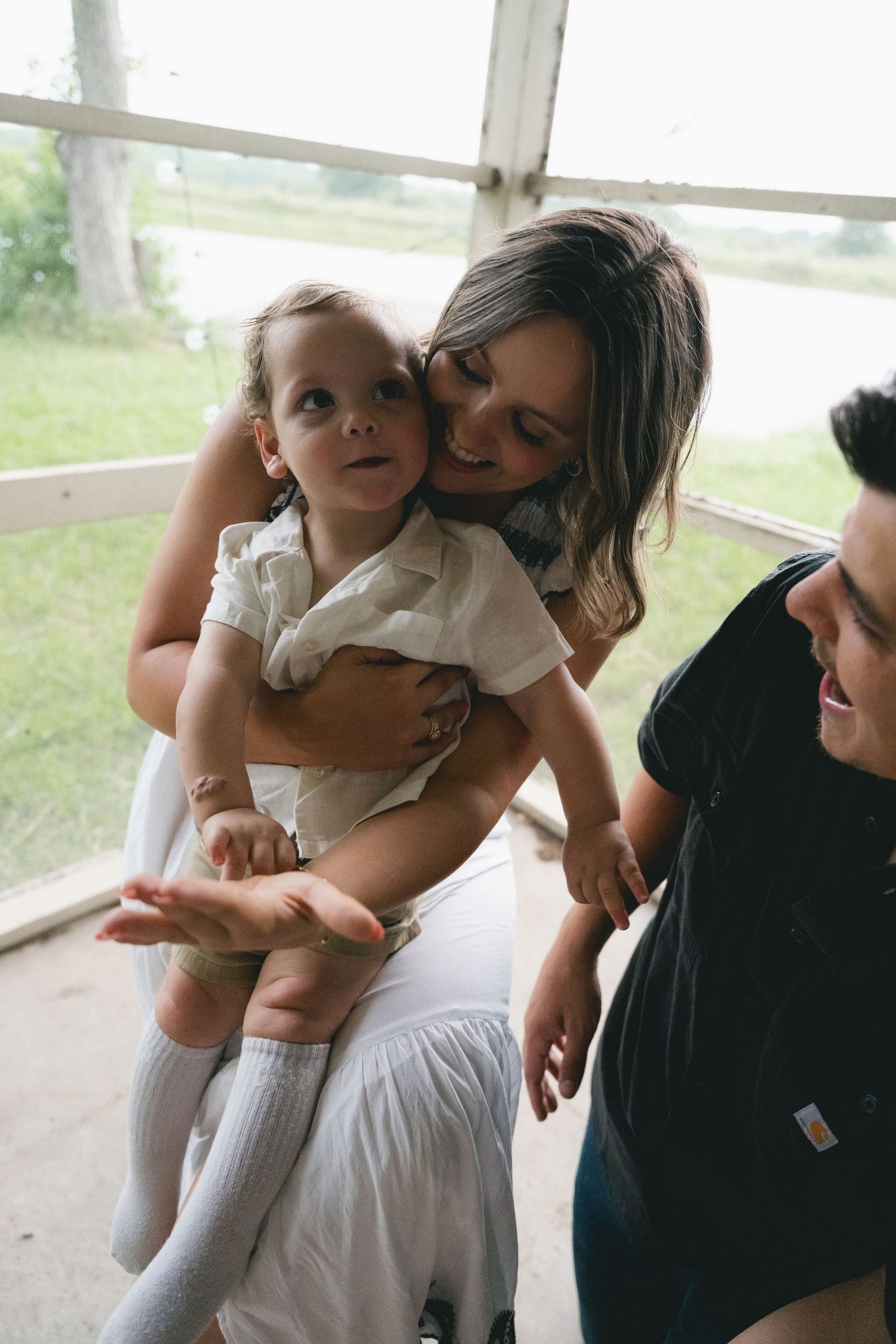 A smiling mother cuddles her toddler while a man looks on inside a bright sunlit room.