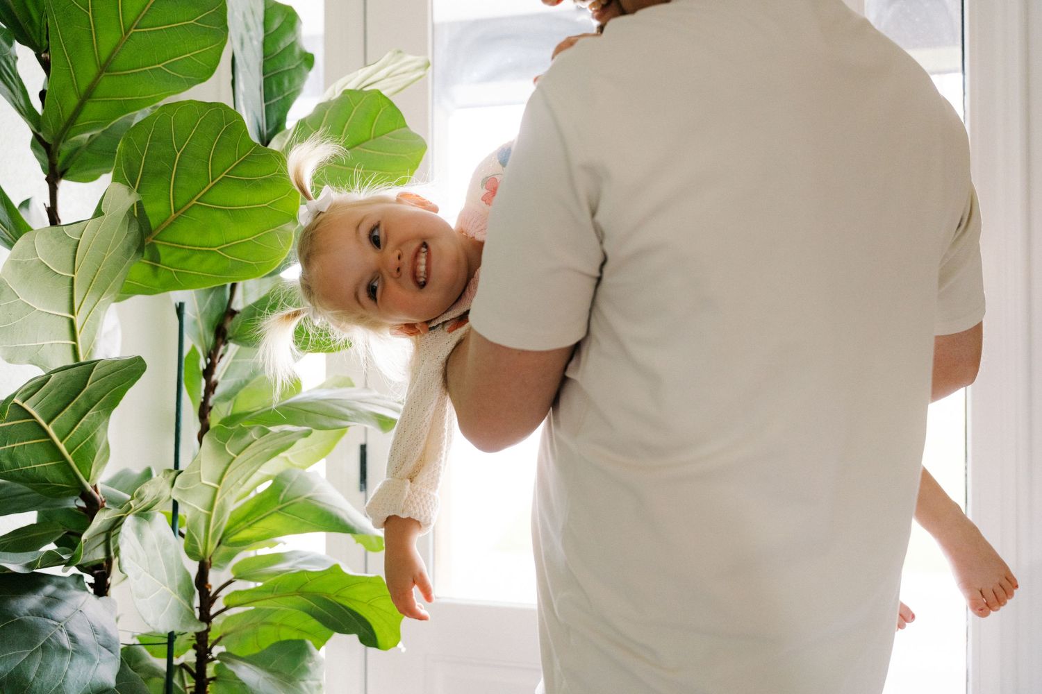 A parent holds a smiling baby upside down playfully near a bright window with green plants.