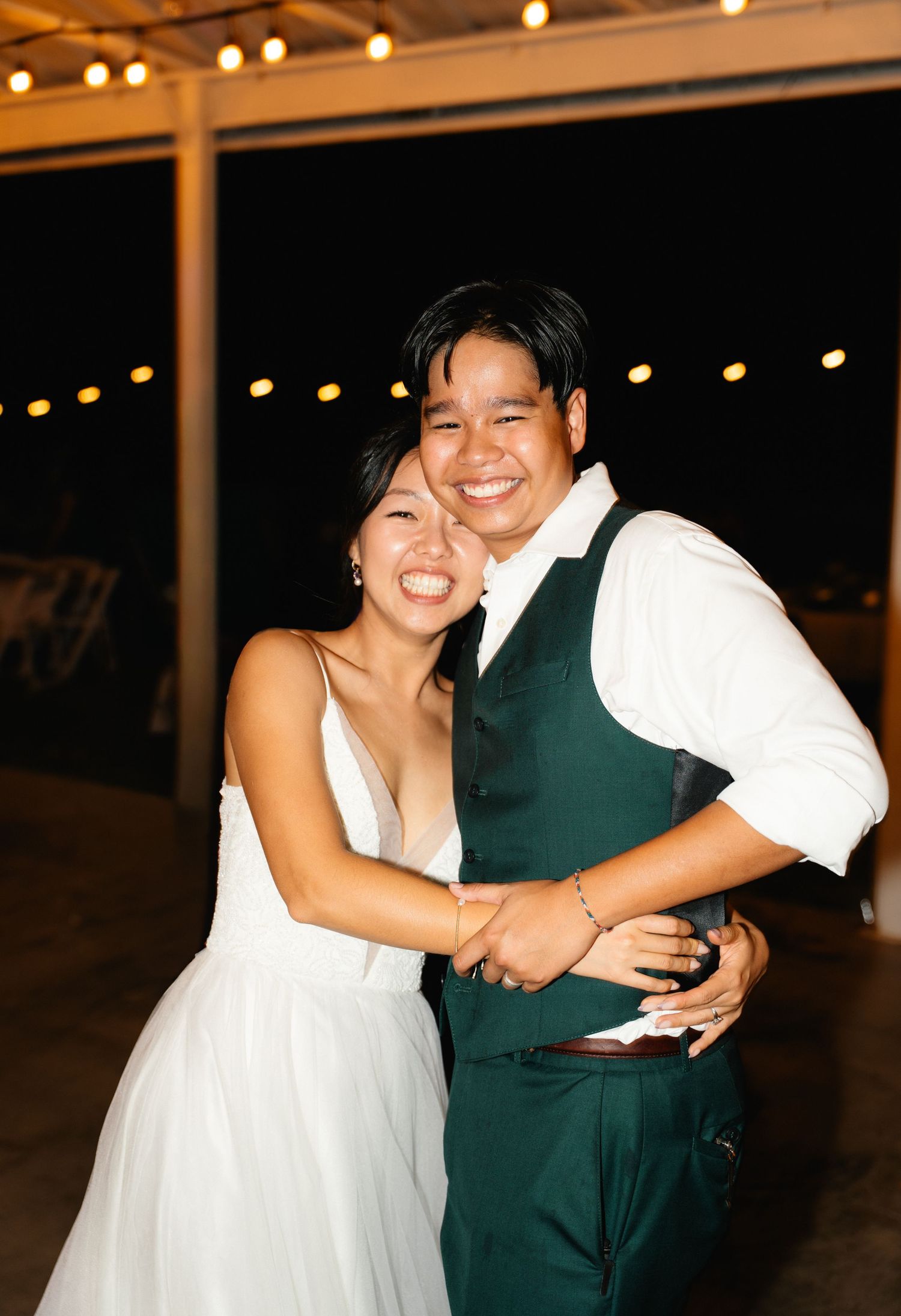 Smiling bride and groom embrace on wedding dance floor at night with string lights glowing behind them.