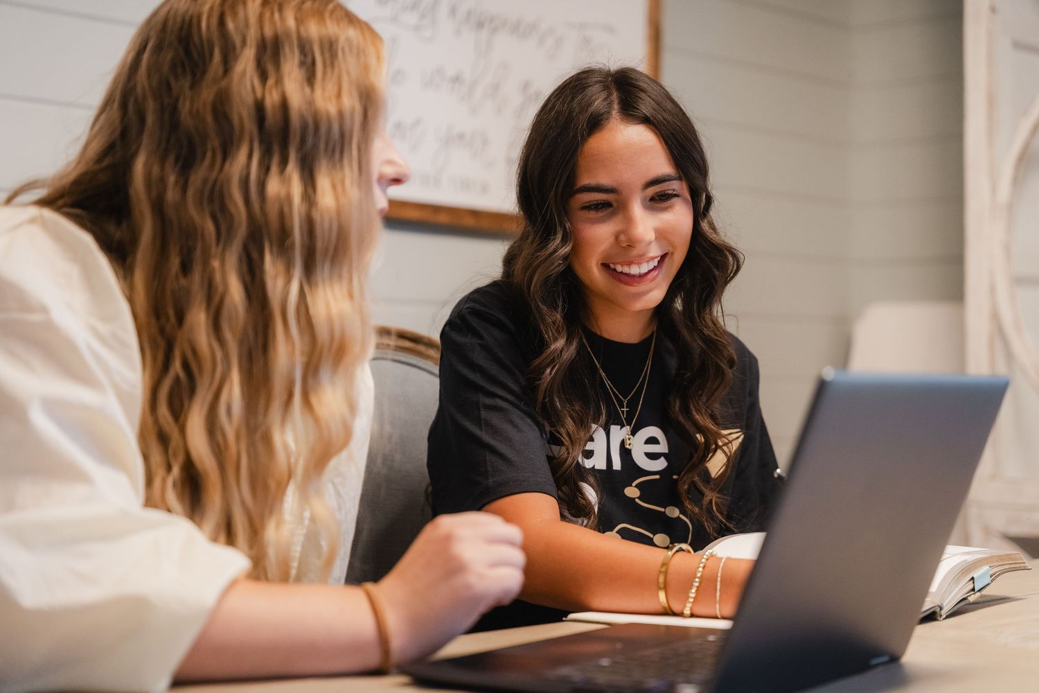 Two young women smiling and collaborating on laptop computer at desk in bright office setting.