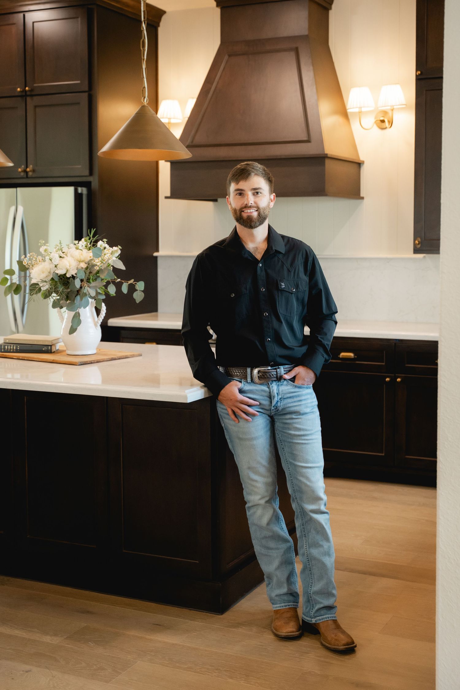 Smiling man in black shirt and jeans leaning against a dark wood kitchen island in an elegant modern kitchen.