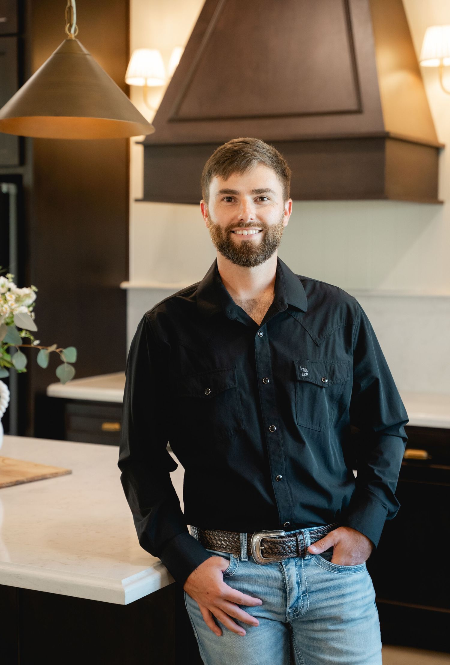 Young man in black western shirt and jeans smiling in a modern dark kitchen with marble countertops.