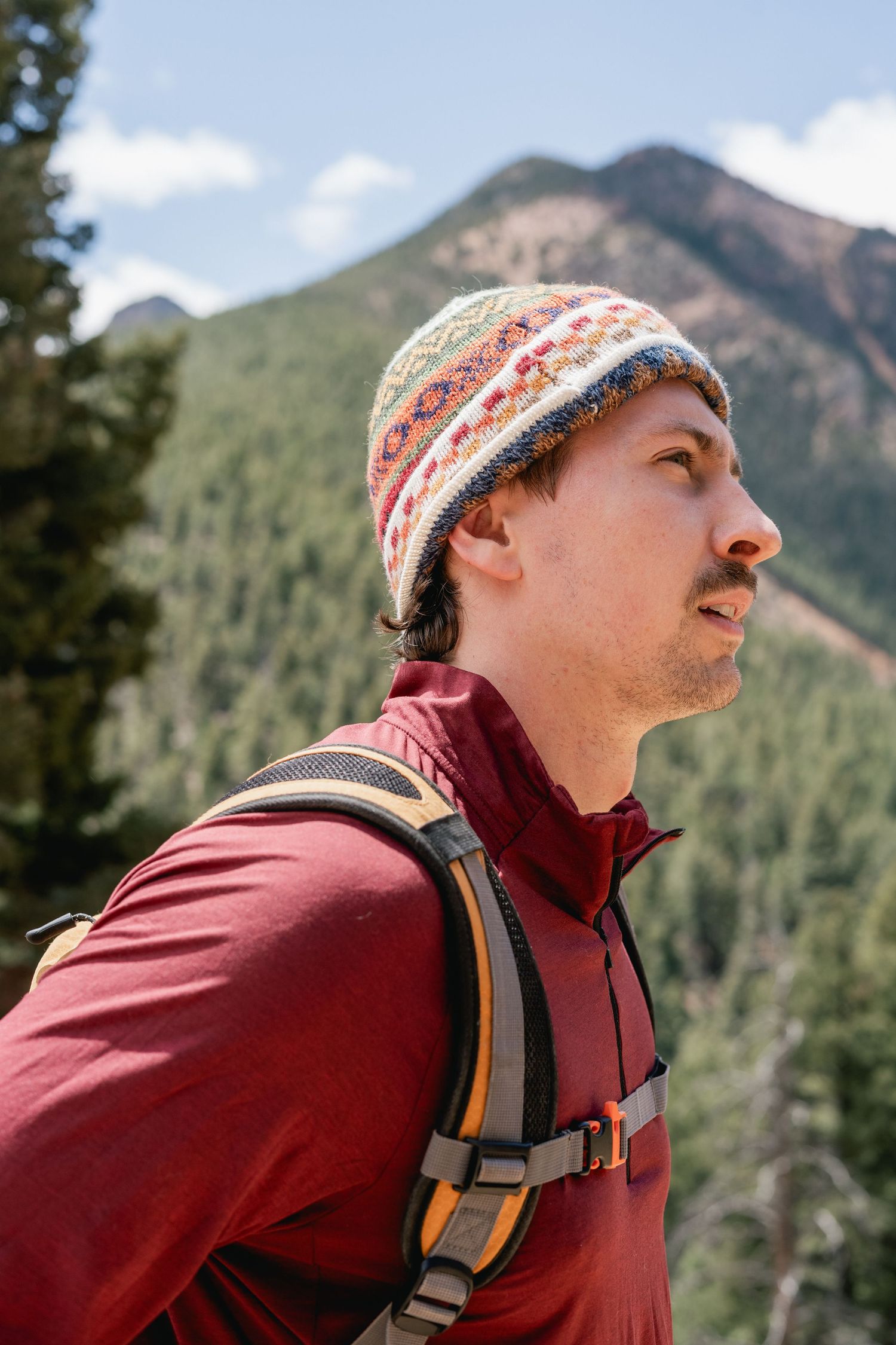 A hiker wearing a colorful knit beanie and red jacket with backpack stands against a mountain forest backdrop.