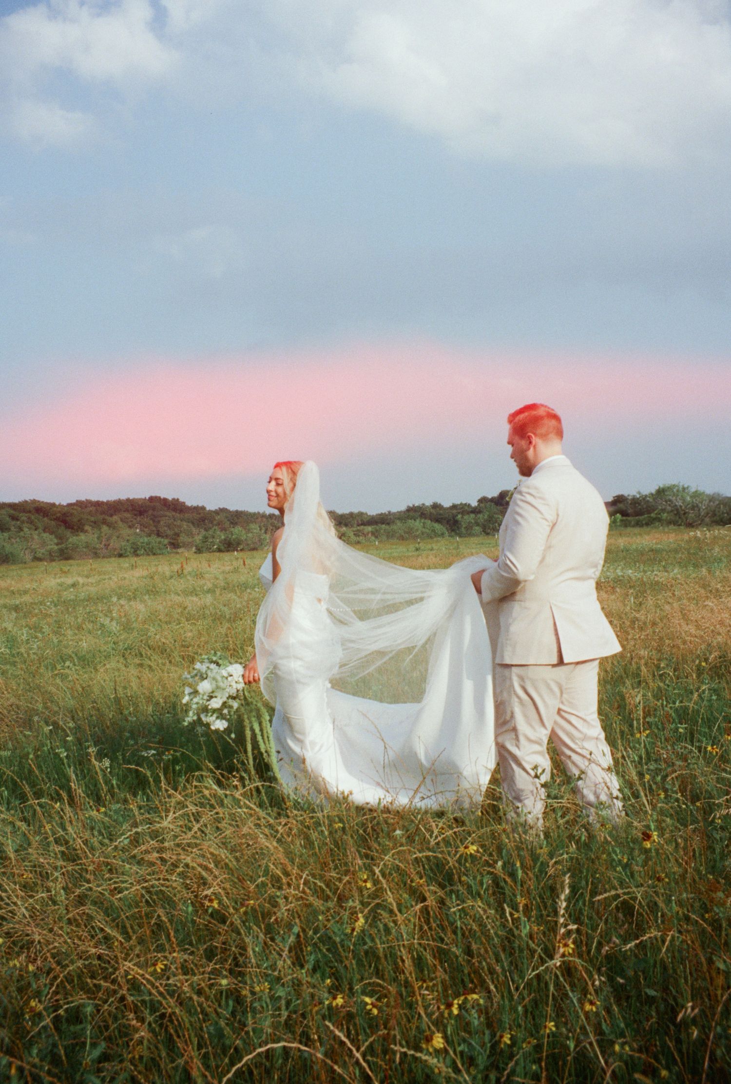 A bride in a white gown and groom in a cream suit stand in a lush green field under a pink-hued sky.