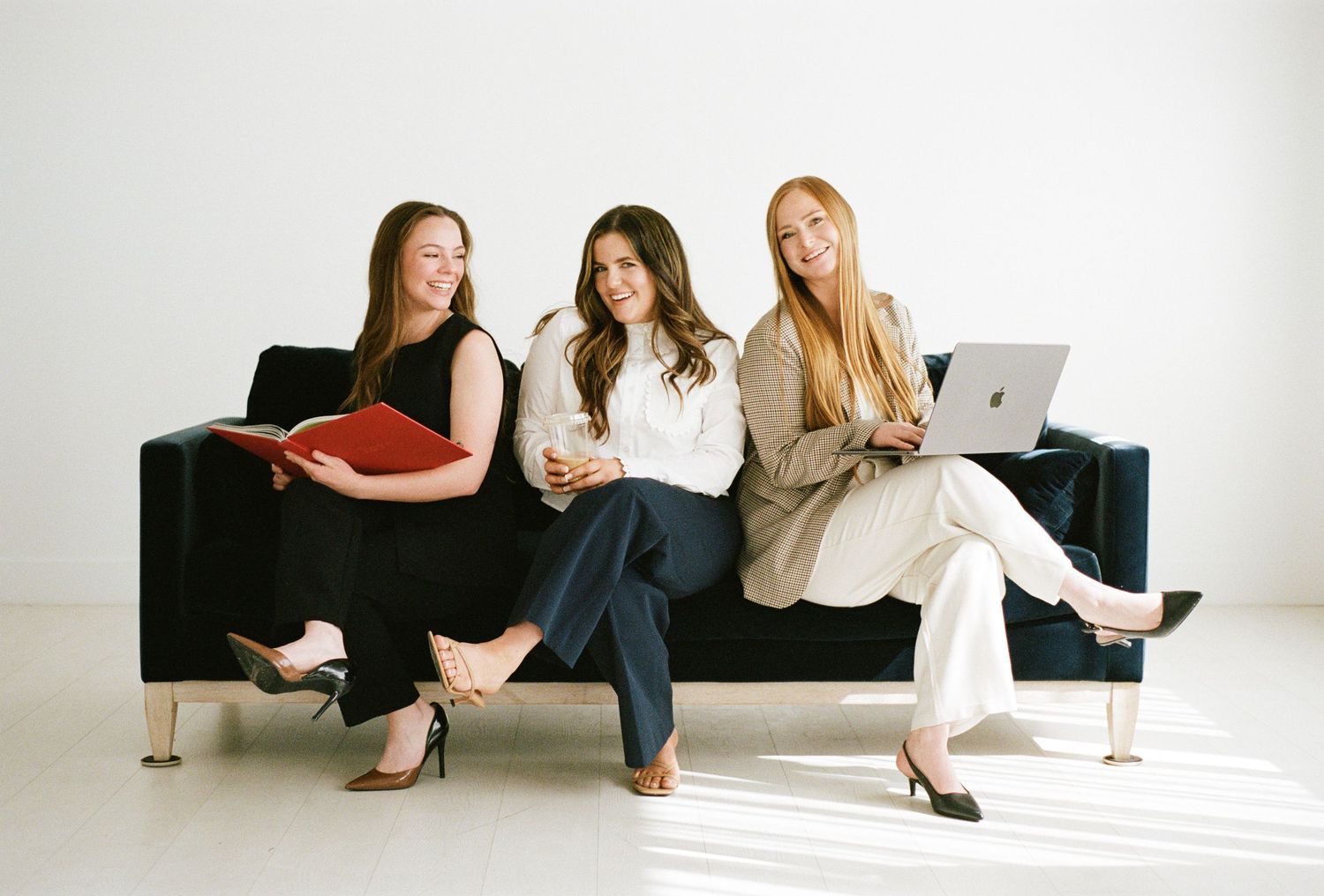 Three professional women sitting on a black sofa, one holding a red folder, one a coffee cup, one a laptop.