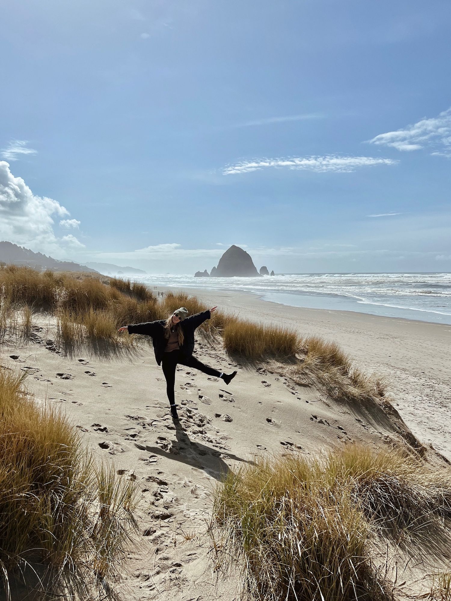 Person posing playfully on sand dunes at Cannon Beach with Haystack Rock in background.