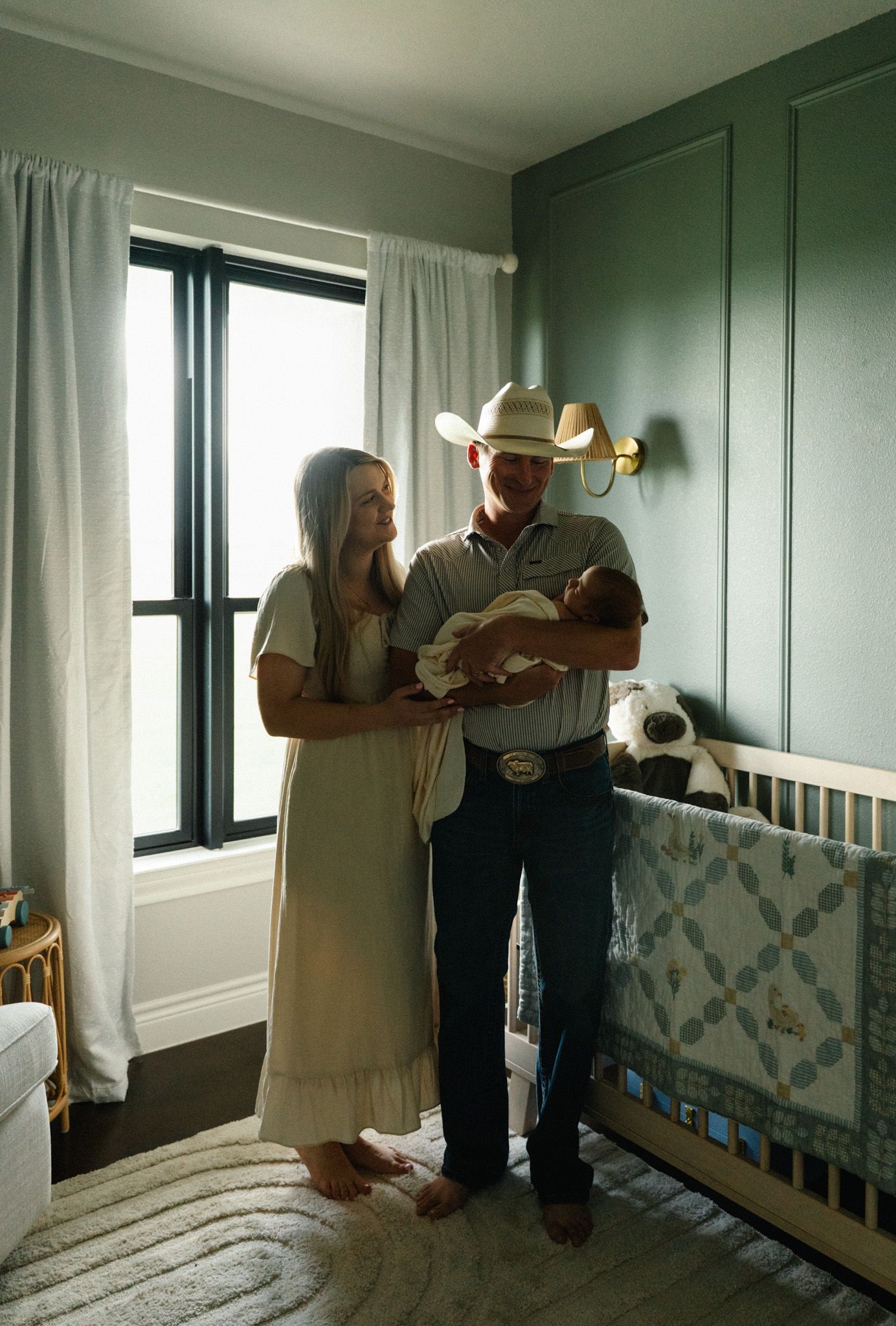 New parents in sage green nursery holding newborn baby beside white crib with quilt.