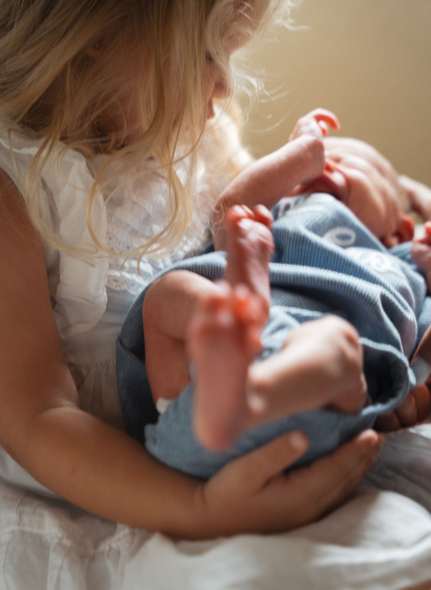 Young blonde girl in white dress carefully holds a sleeping newborn baby.