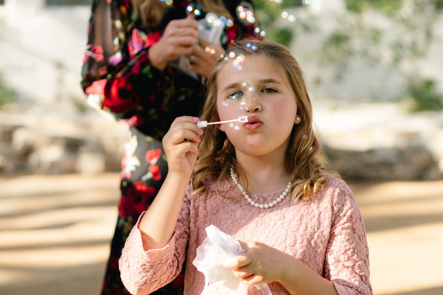 Young girl in pink lace dress and pearl necklace blowing bubbles outdoors at a wedding celebration.