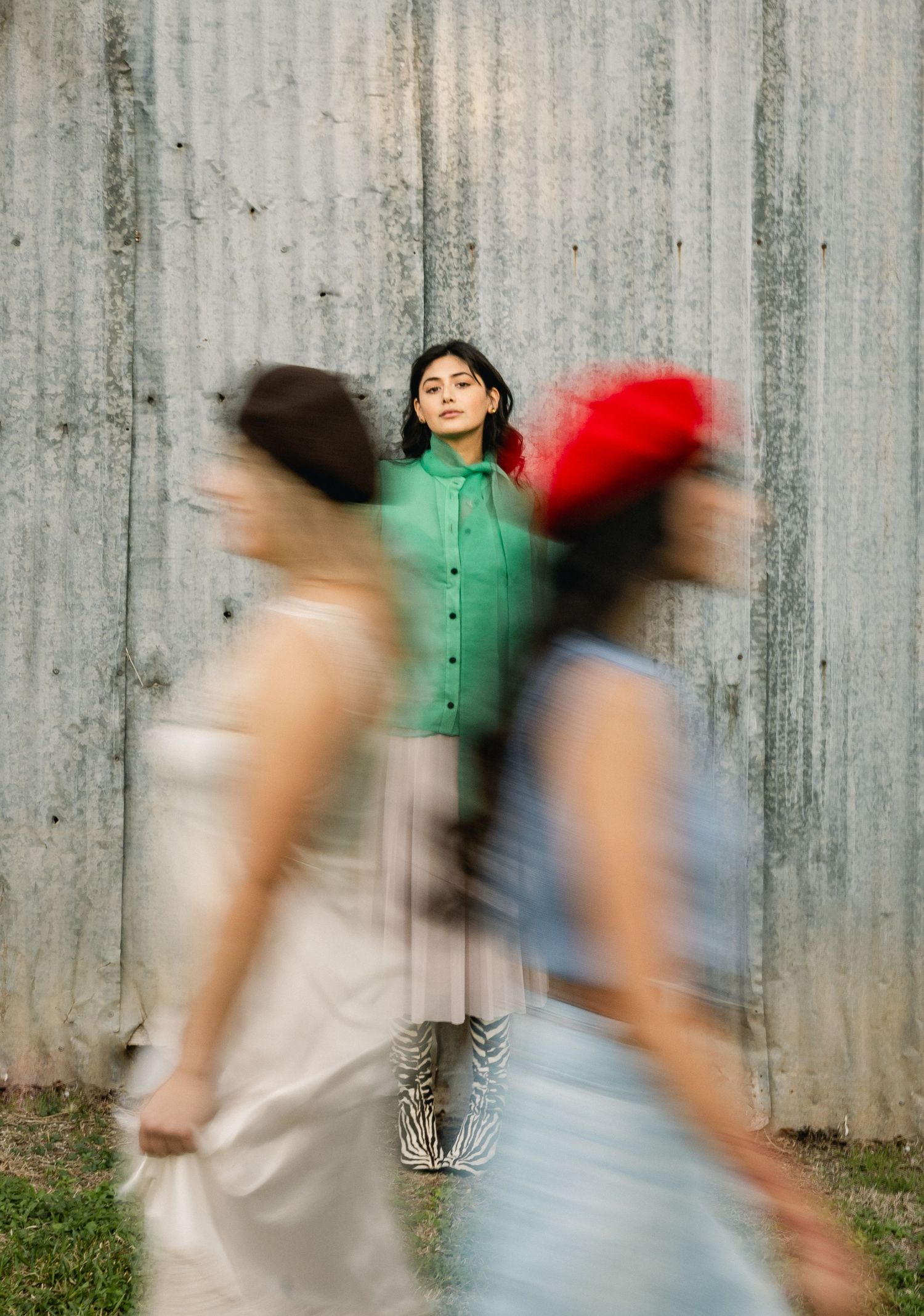 Woman in green shirt standing still against metal wall while two blurred figures walk past.