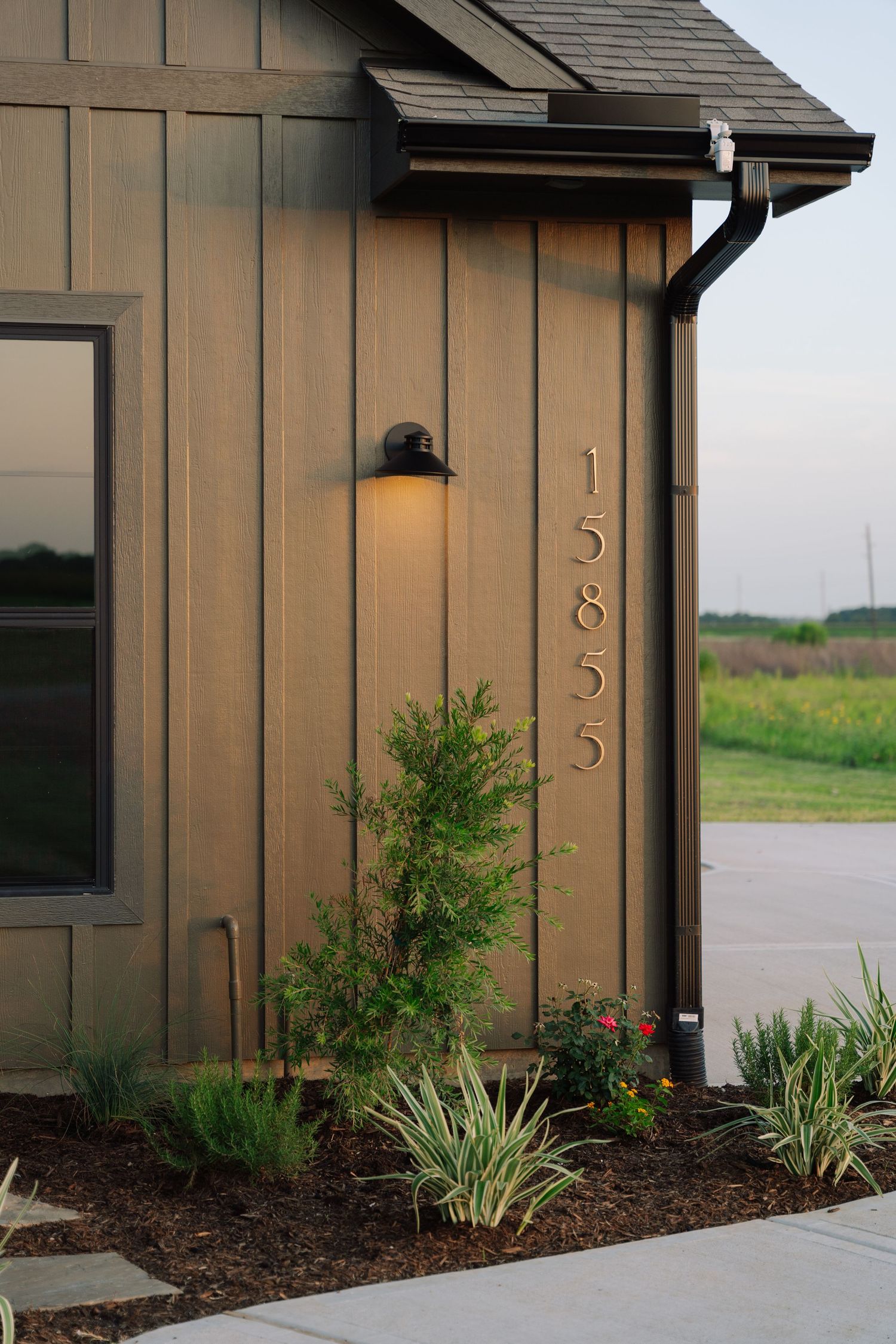 Board and batten exterior siding with vertical house numbers, wall sconce, and manicured landscaping at dusk.