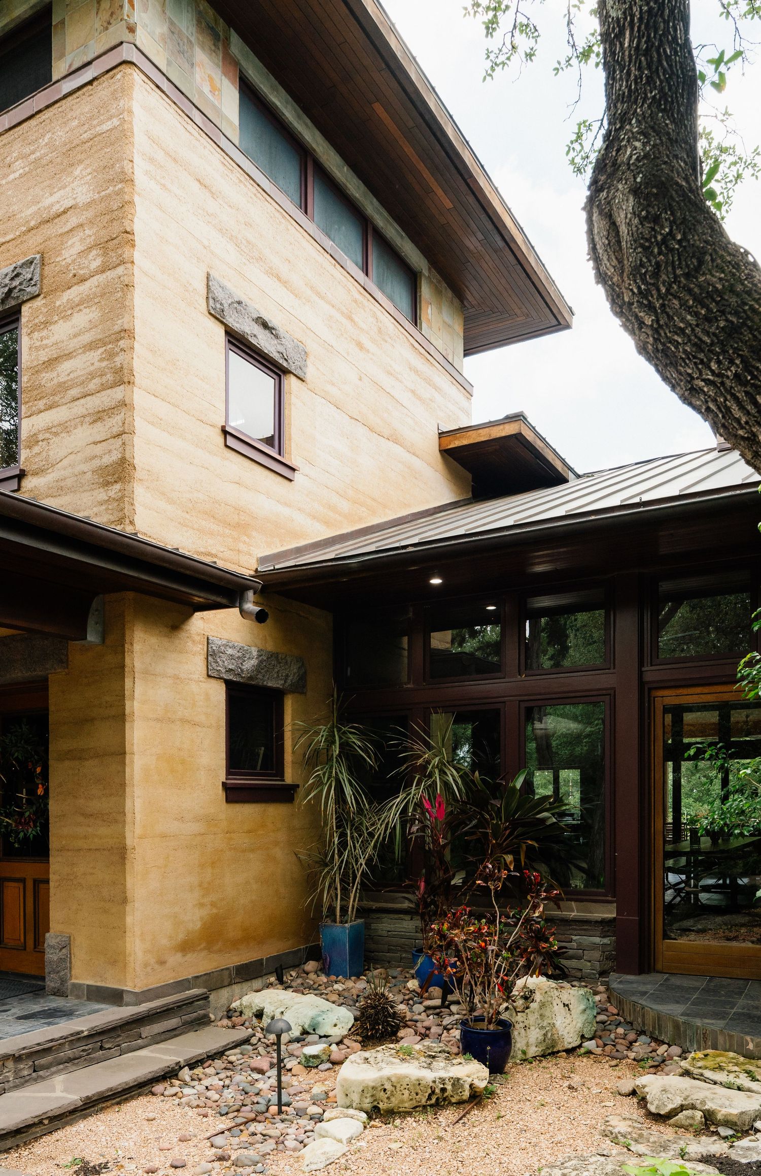 Two-story rammed earth home with metal roof, dark wood trim, and desert landscaping with rocks and plants.