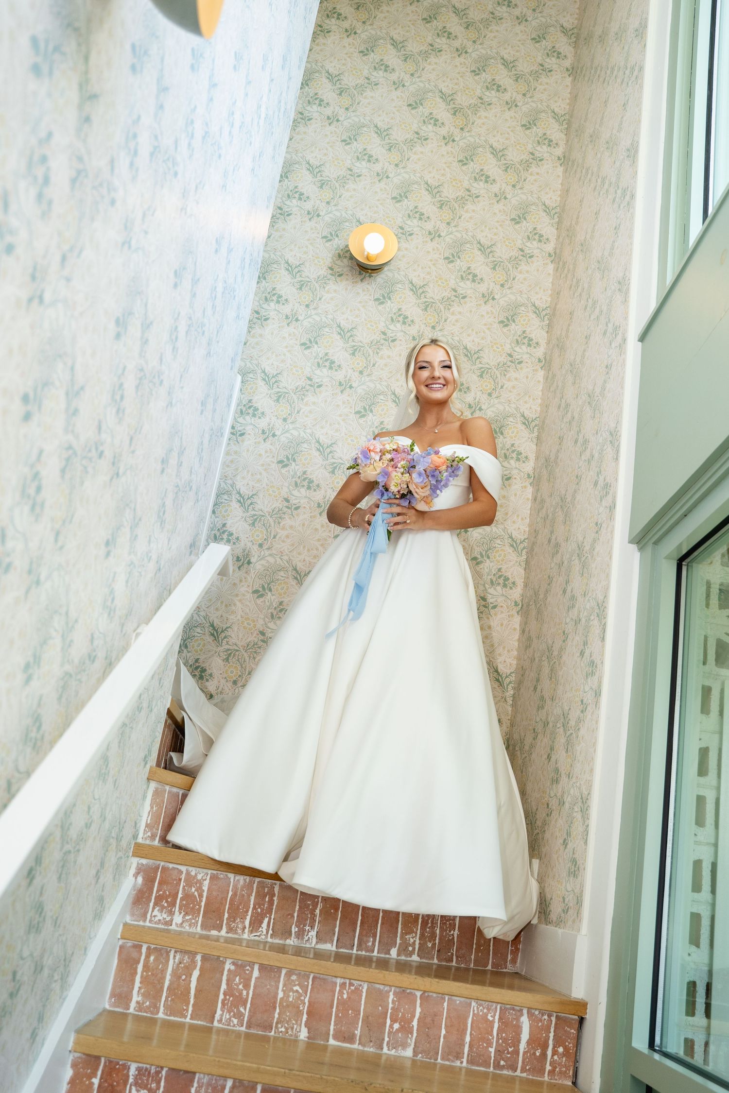 Smiling bride in off-shoulder ball gown holds colorful bouquet while standing on staircase with floral wallpaper.