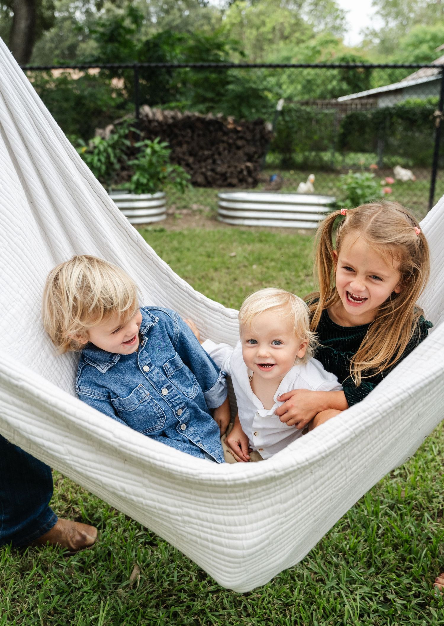 Three children laugh and cuddle together in a white hammock in a lush backyard garden.