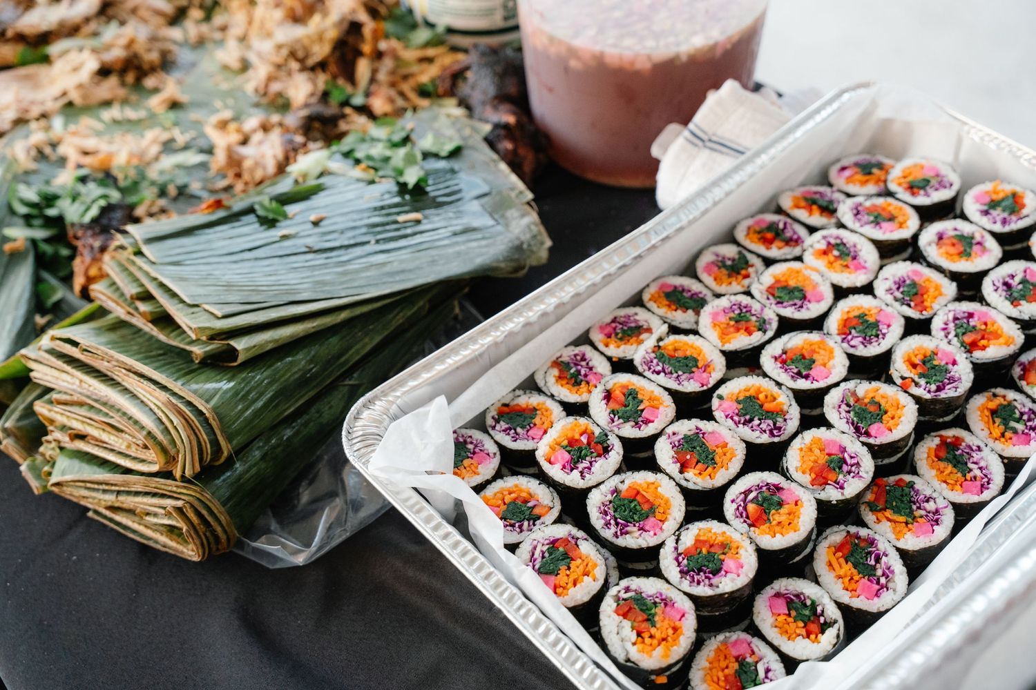 A tray of colorful sushi rolls displayed alongside wrapped tamales at a wedding reception.