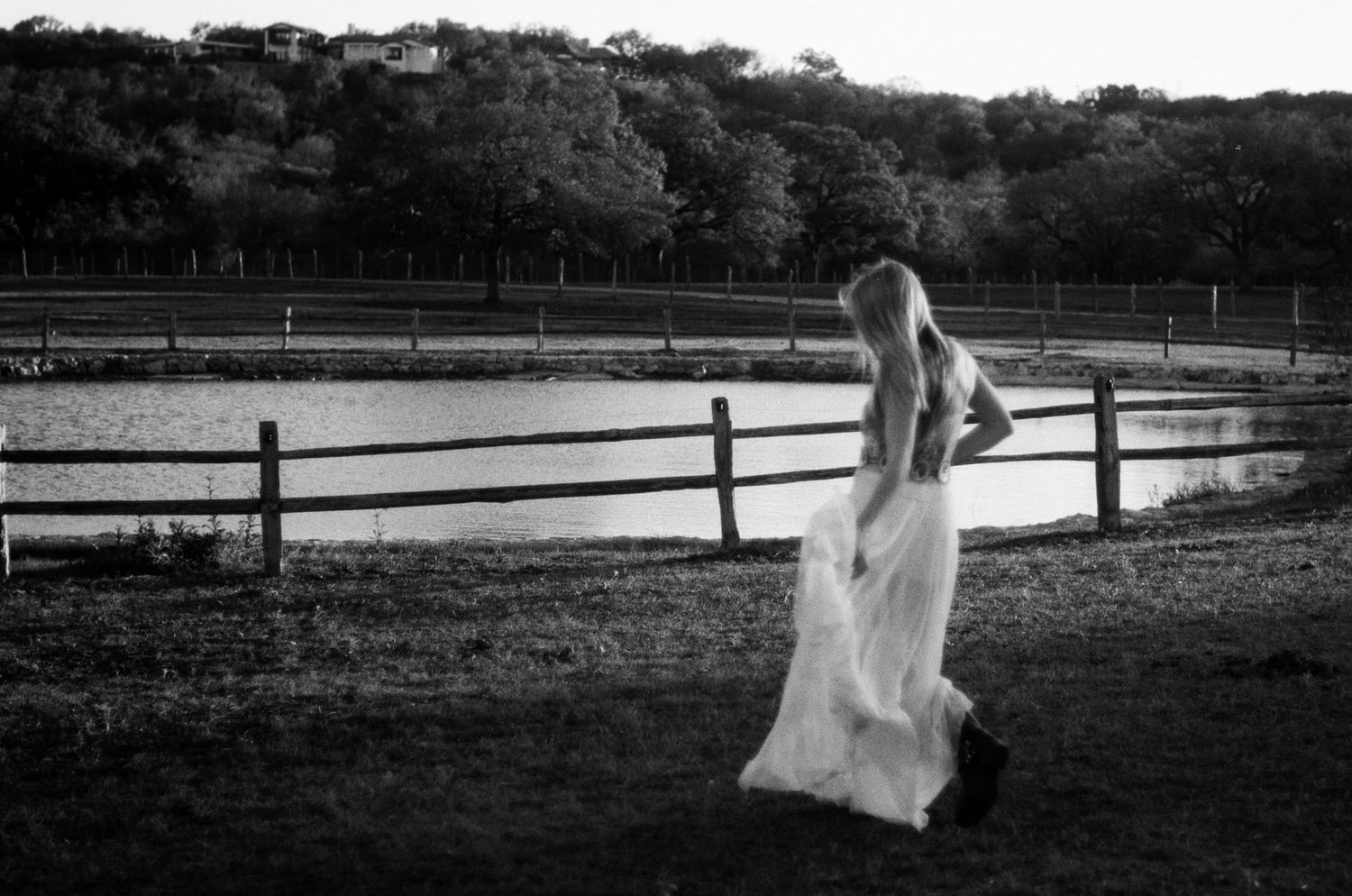 Black and white photo of a bride in a flowing white gown standing by a wooden fence near a pond.
