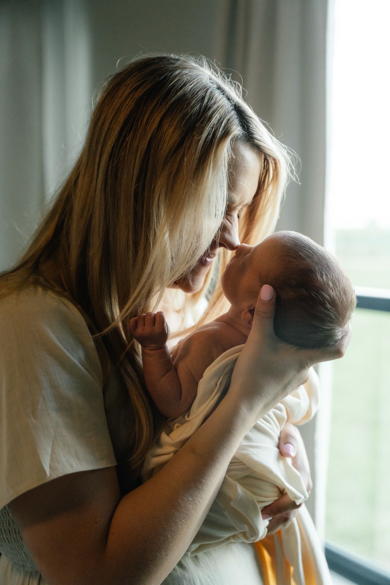 Blonde mother gently touching her nose to her swaddled newborn baby's head near a window.
