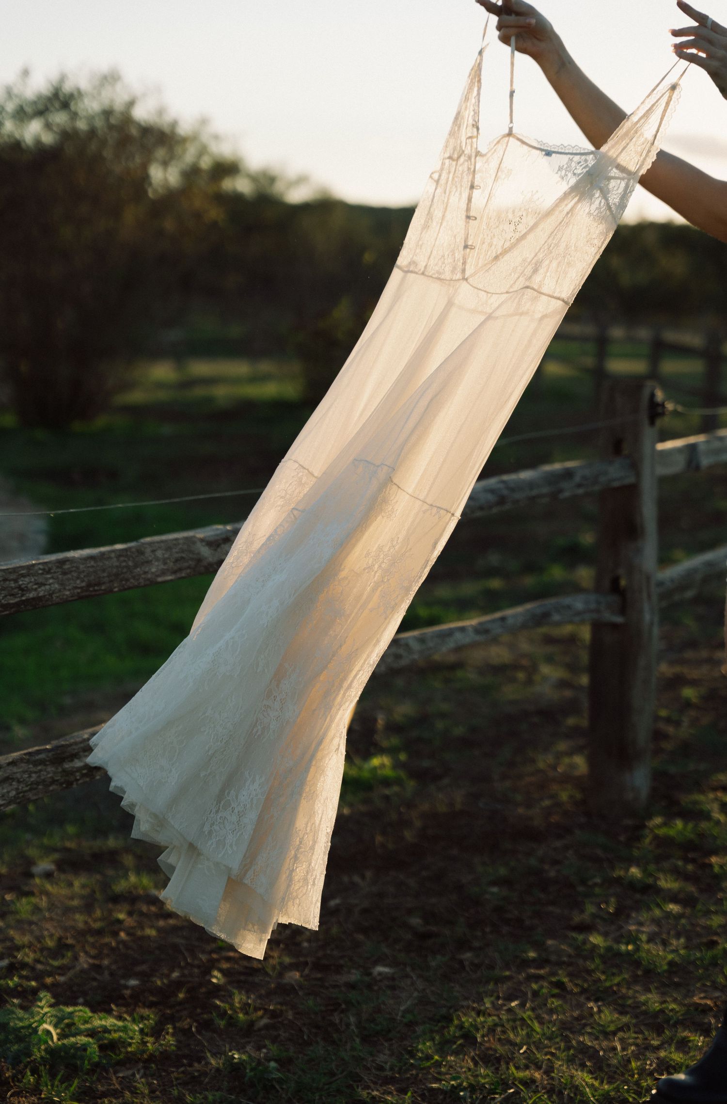 Sheer white lace dress held up on rustic wooden fence at golden hour sunset.