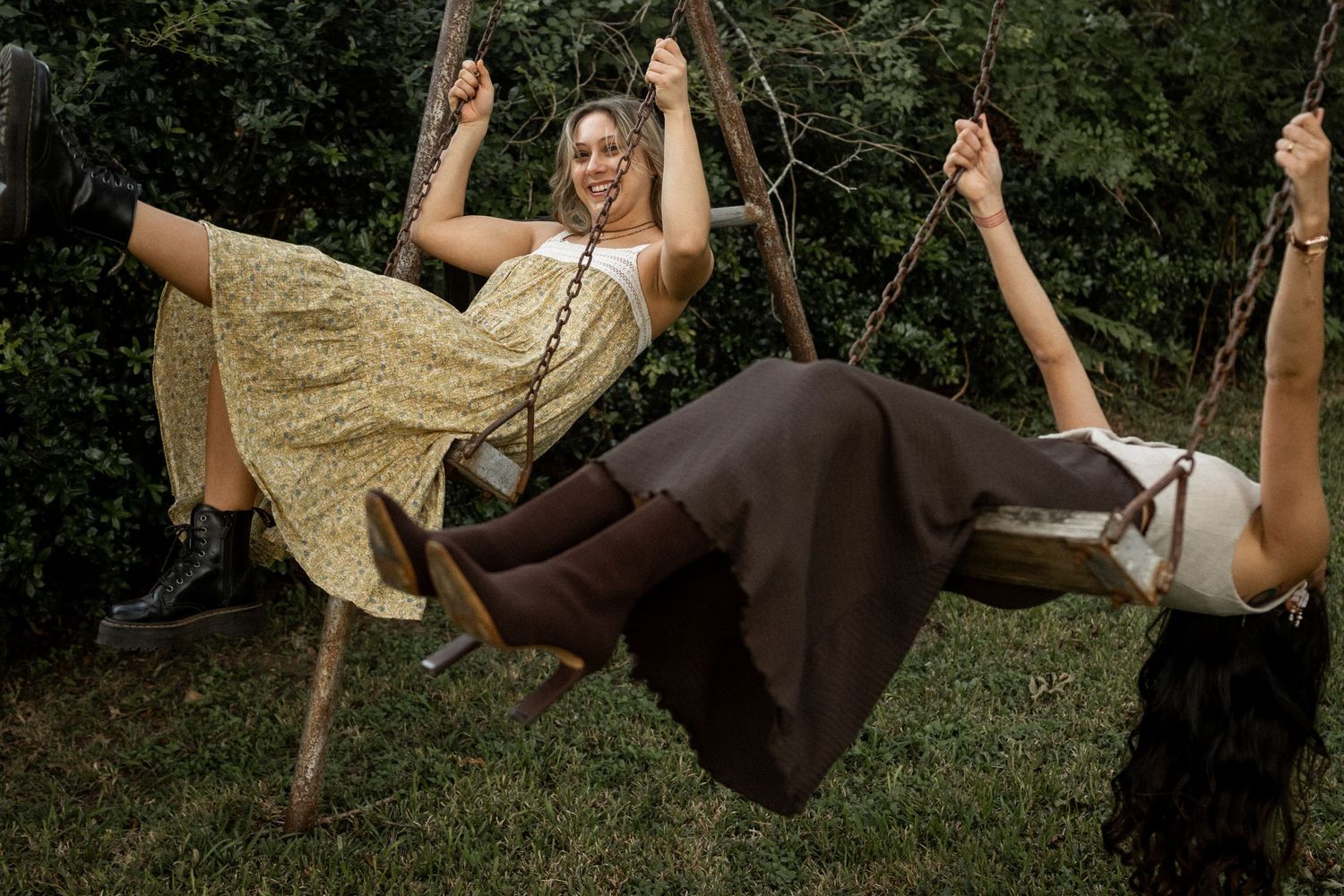Two women in vintage dresses swing playfully on a wooden swing set outdoors in a lush green garden.