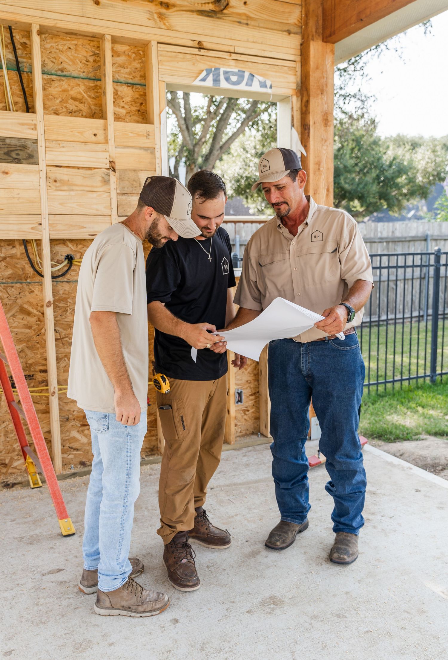 Three construction workers reviewing building plans at an outdoor framed structure with a red ladder nearby.