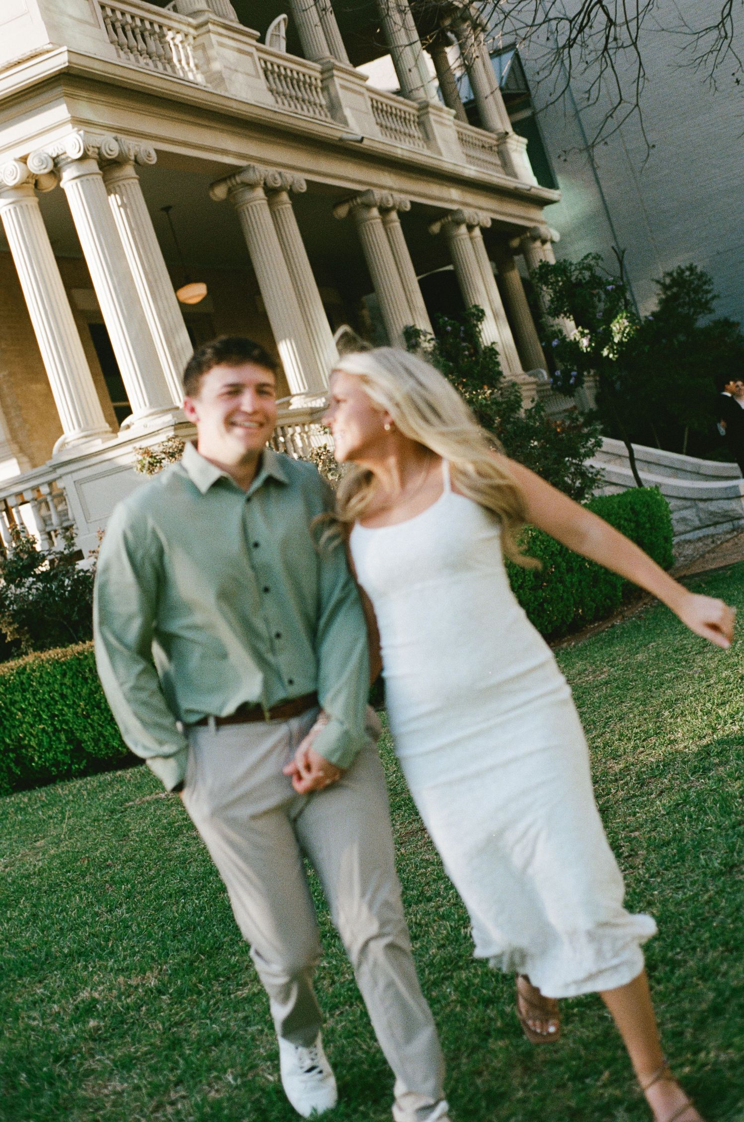 Joyful couple holding hands and laughing while walking on grass in front of columned estate.