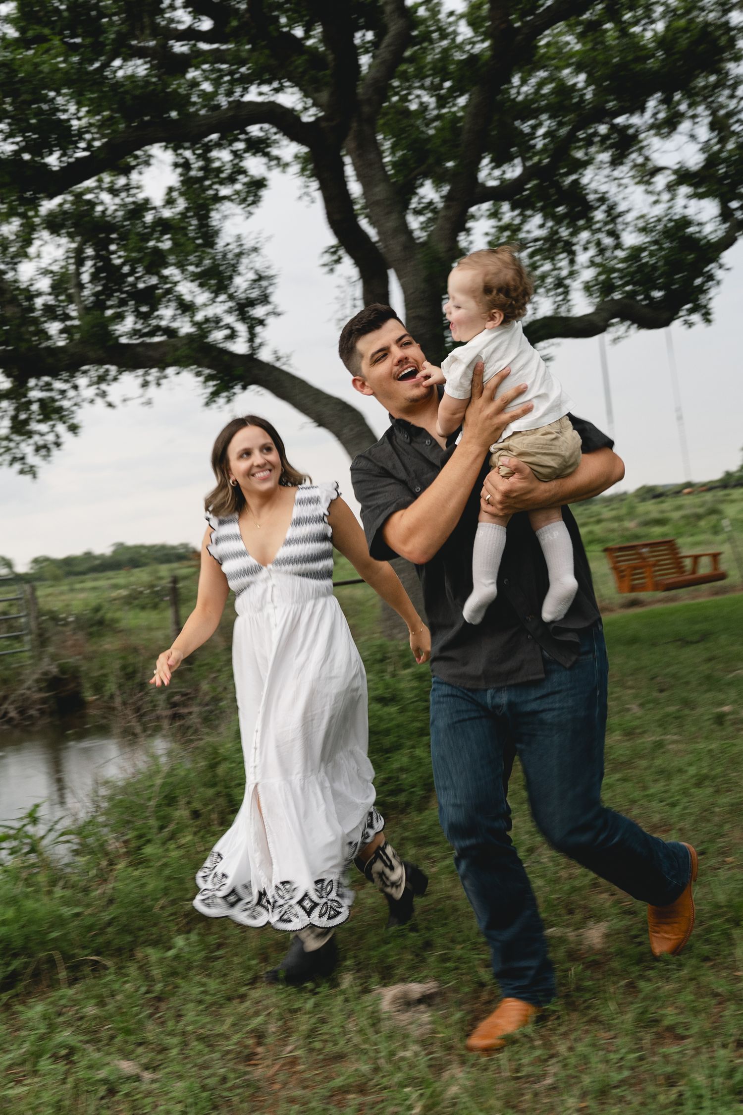 A joyful family runs outdoors near a pond, with father laughing while holding their baby and mother smiling beside them.