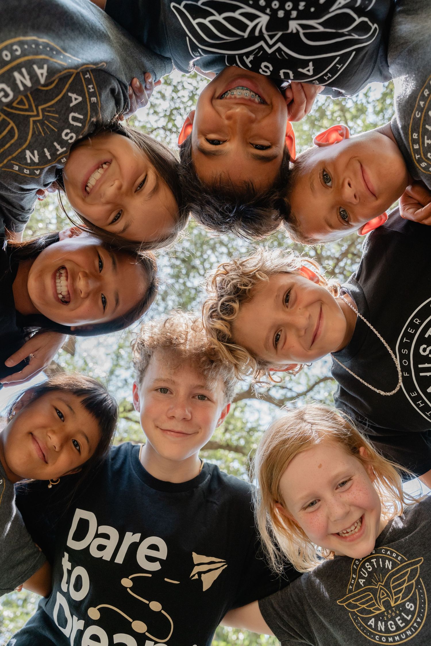 Seven smiling children in black Austin Angels shirts huddle together looking down at camera outdoors.