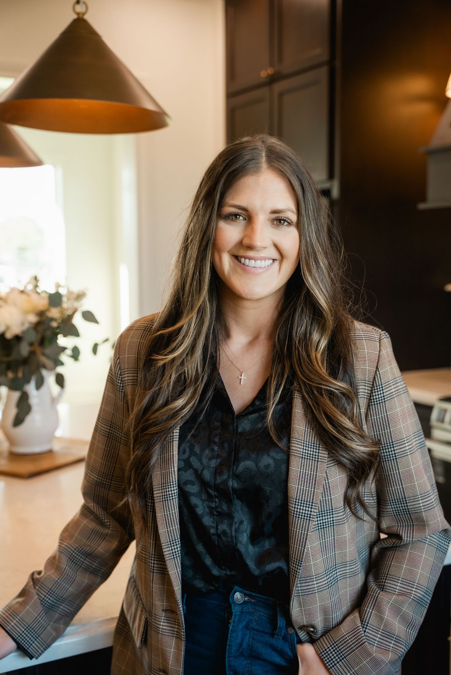 Smiling woman with long brunette hair wearing a plaid blazer poses in a modern kitchen interior.