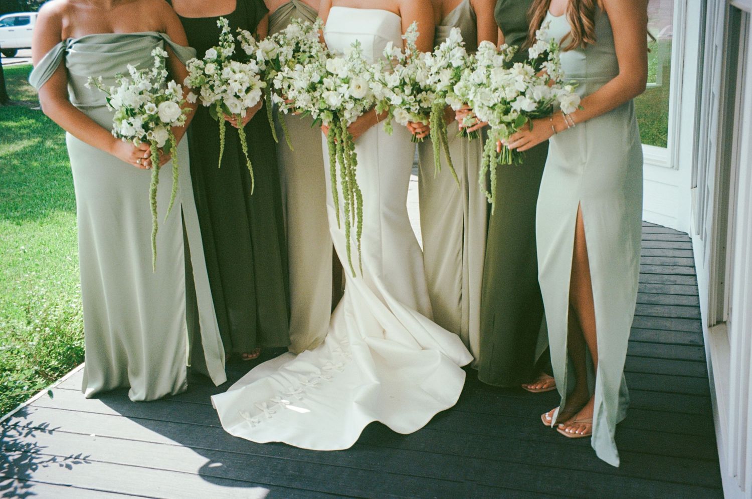 Bride in white gown with bridesmaids in sage green dresses holding white floral bouquets on a porch.