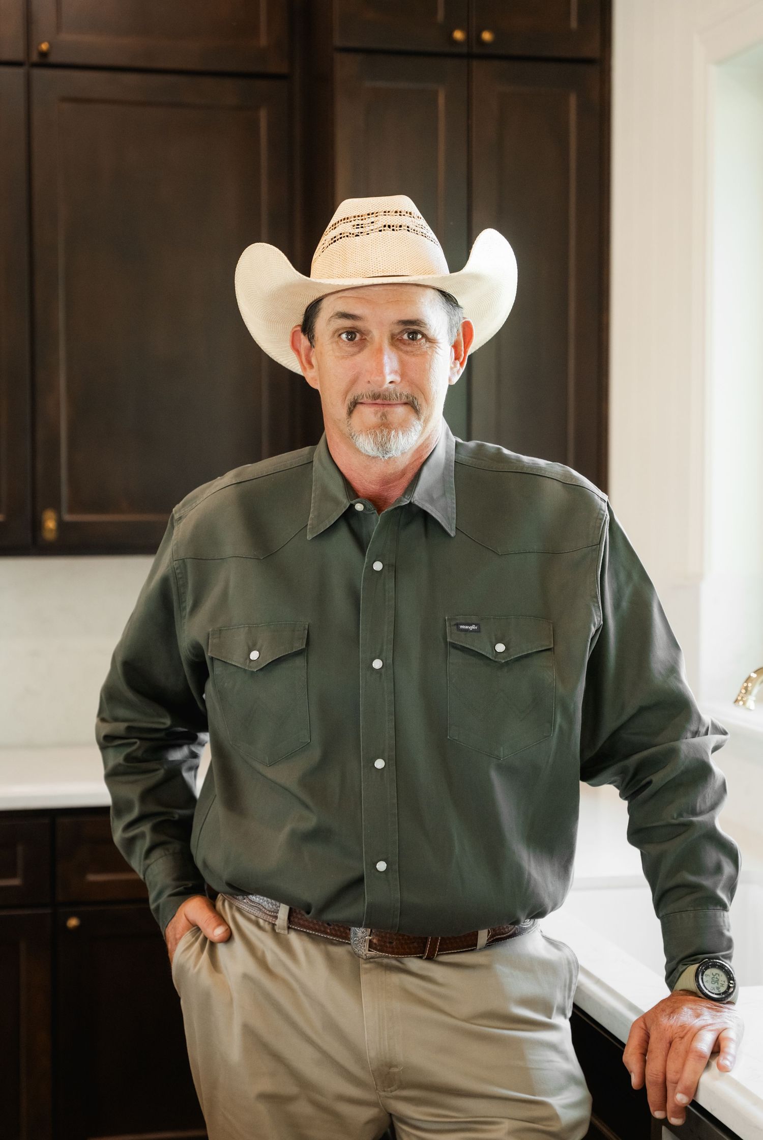 Man in white cowboy hat and green western shirt leaning against white kitchen counter with dark wood cabinets.