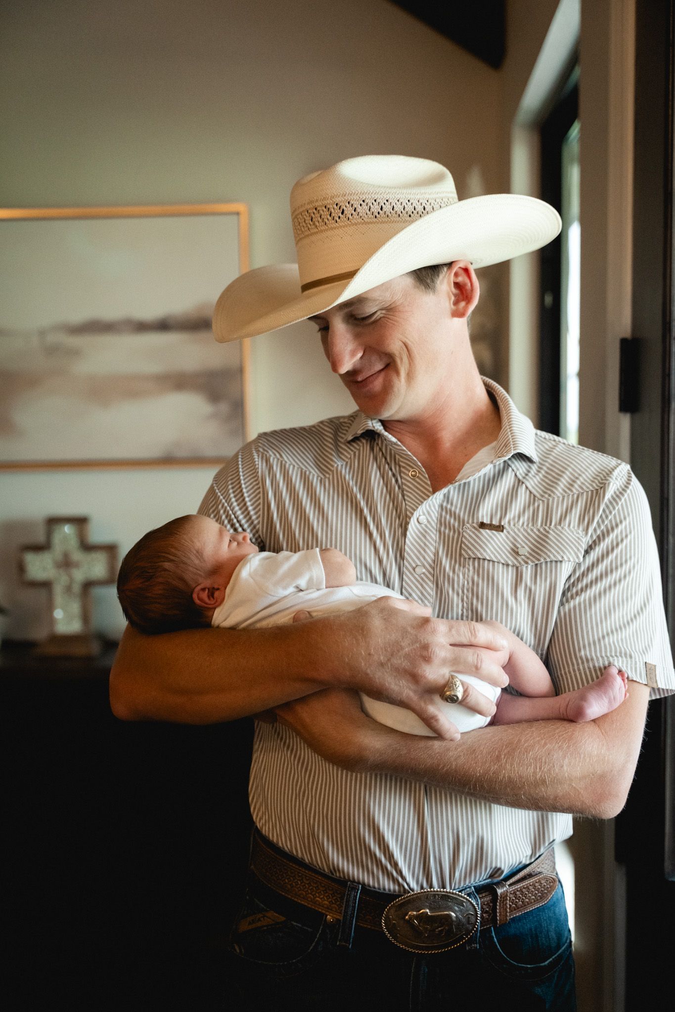 Proud father in cowboy hat and striped shirt gently cradling his newborn baby at home.