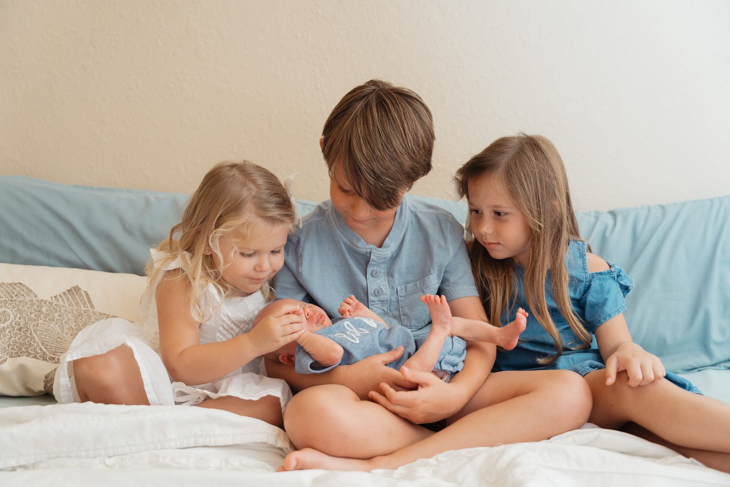 Three siblings sit together on a bed, gently touching their newborn baby sibling.