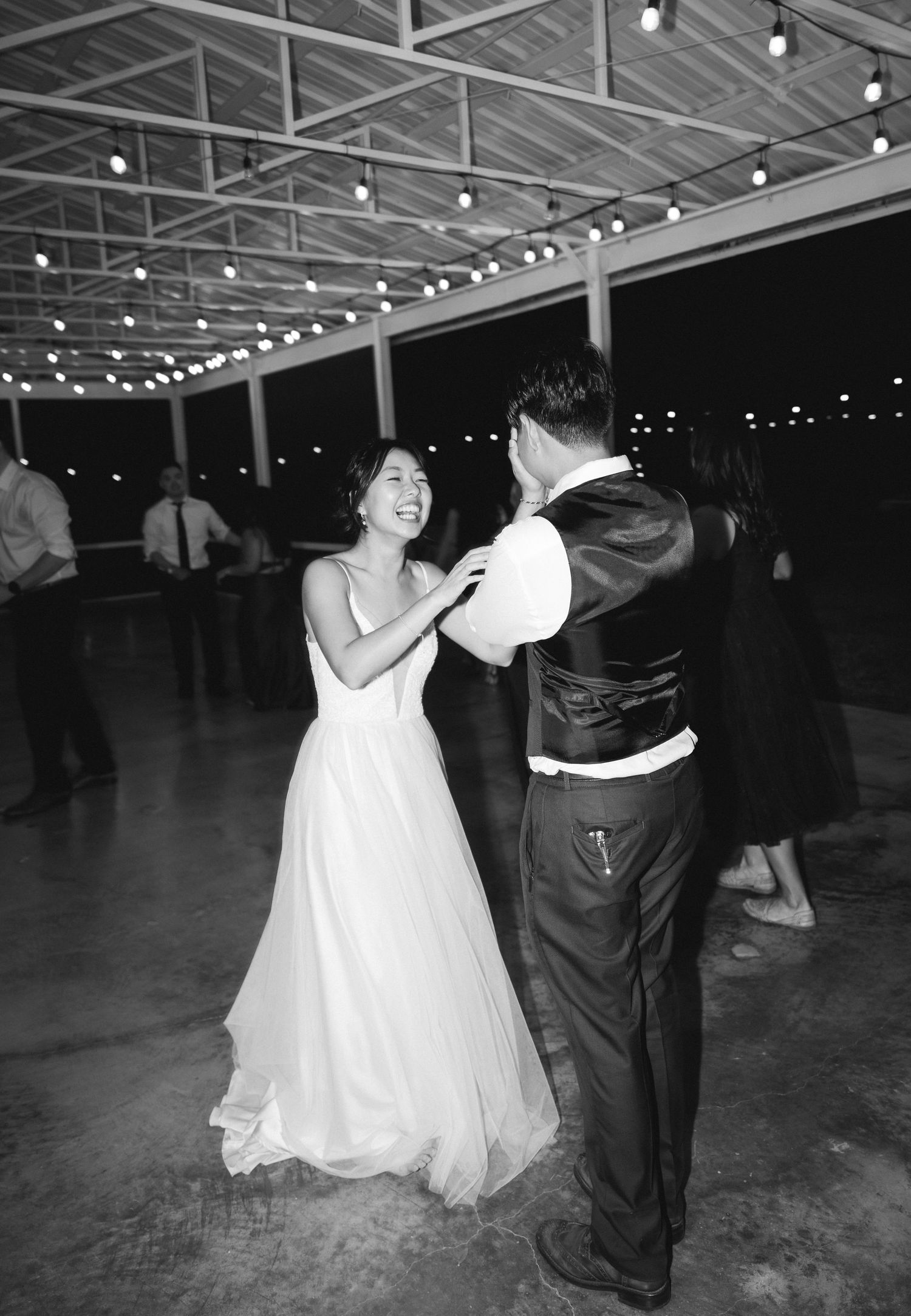Bride and groom share a romantic first dance under string lights in black and white.