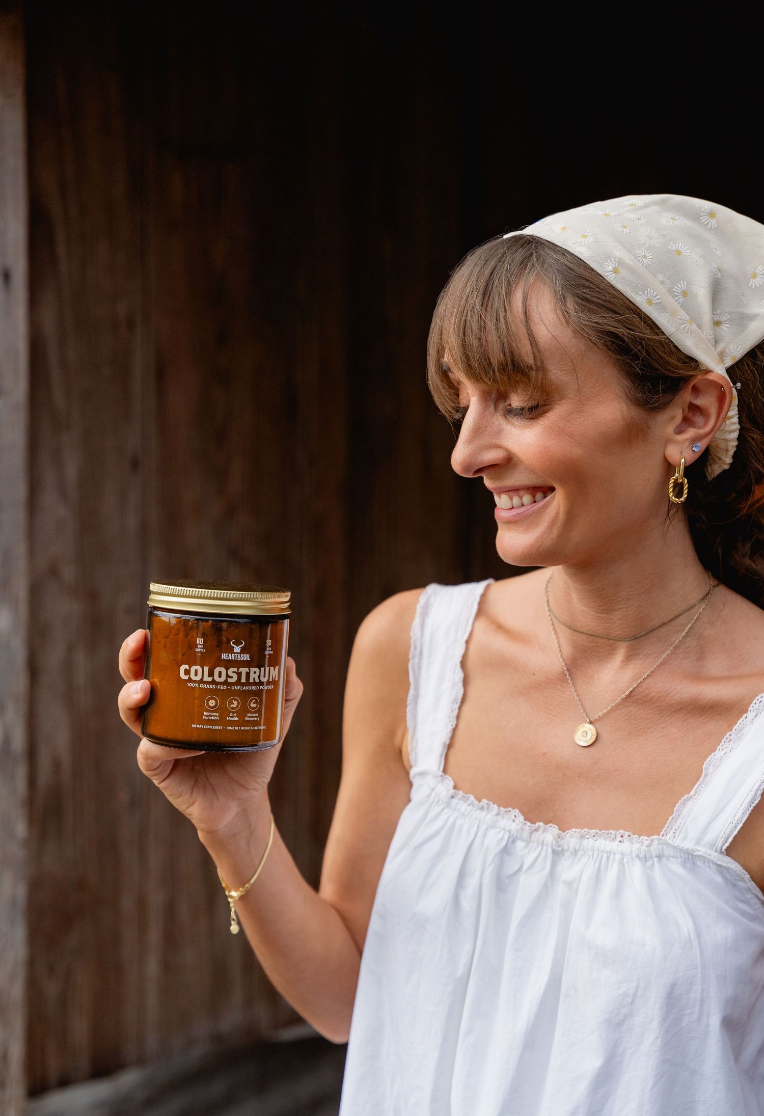 Smiling woman in white dress holding a jar of Colostrum supplement outdoors.