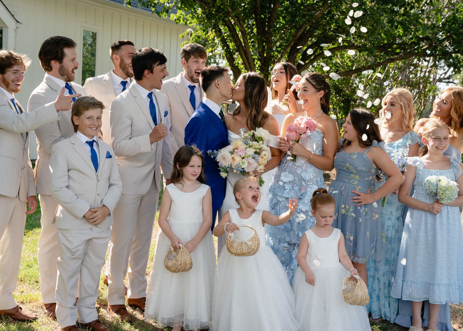 Large wedding party with bridesmaids in blue dresses and groomsmen in tan suits with flower girls.
