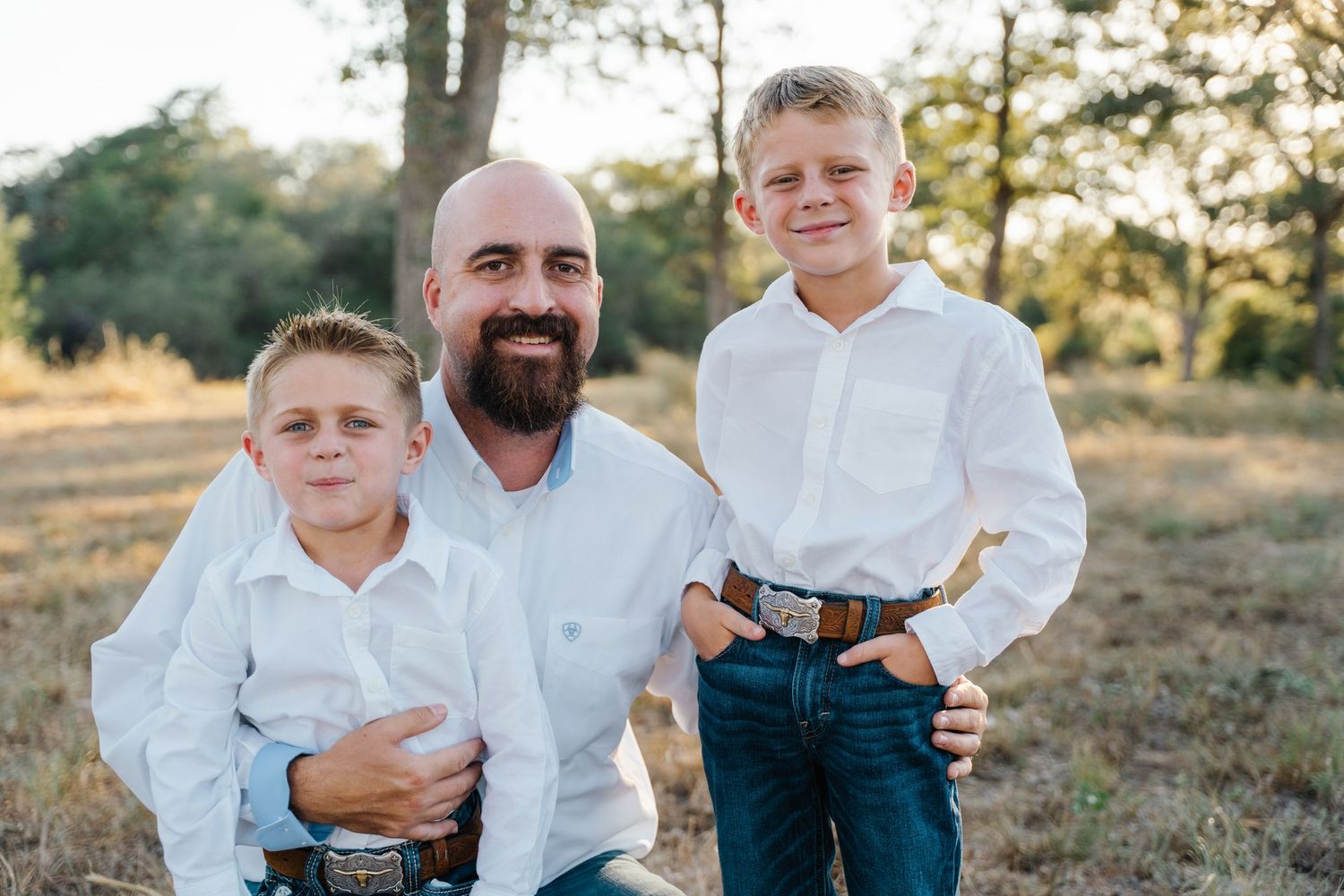 A bald bearded father smiles proudly with his two sons in a sunny field.