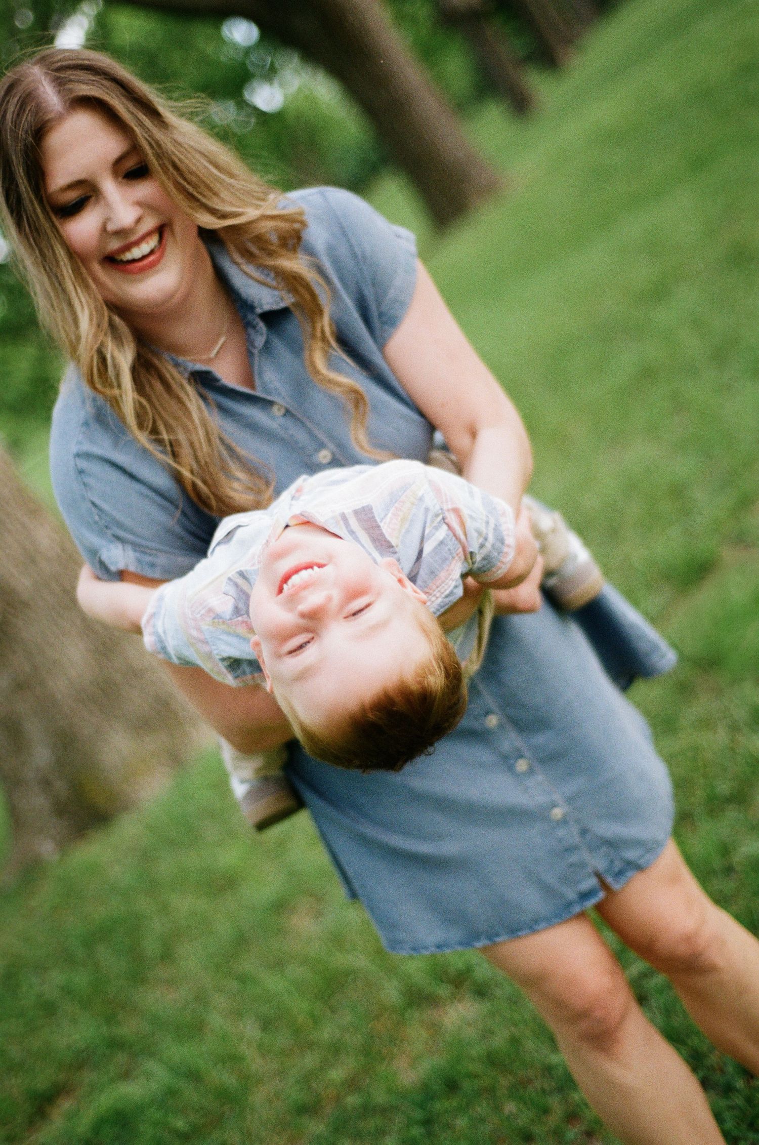 A laughing mother holds her giggling child upside down in a lush green park.
