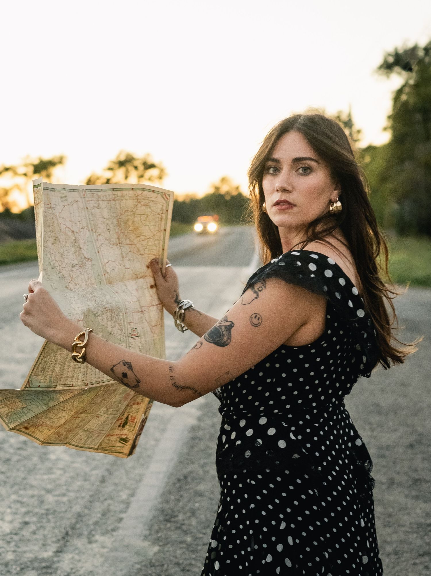 Woman in polka dot dress holding vintage map on roadside at golden hour, showcasing arm tattoos and gold jewelry.