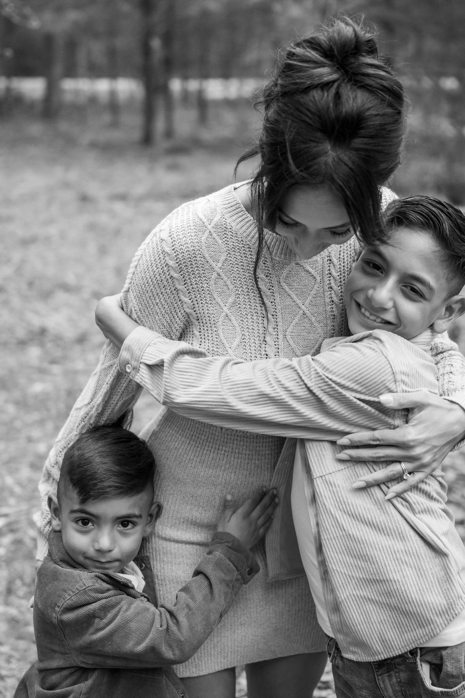 Black and white portrait of a mother embracing her two young sons in a warm outdoor family moment.