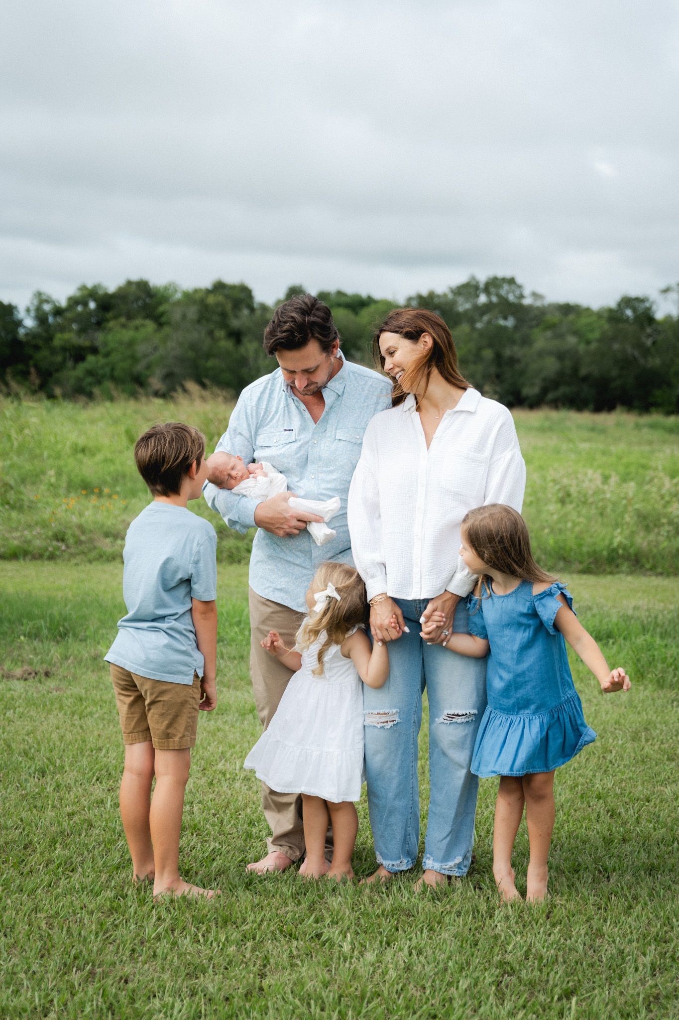 Happy family of six standing in a green field, parents holding newborn surrounded by three young children.