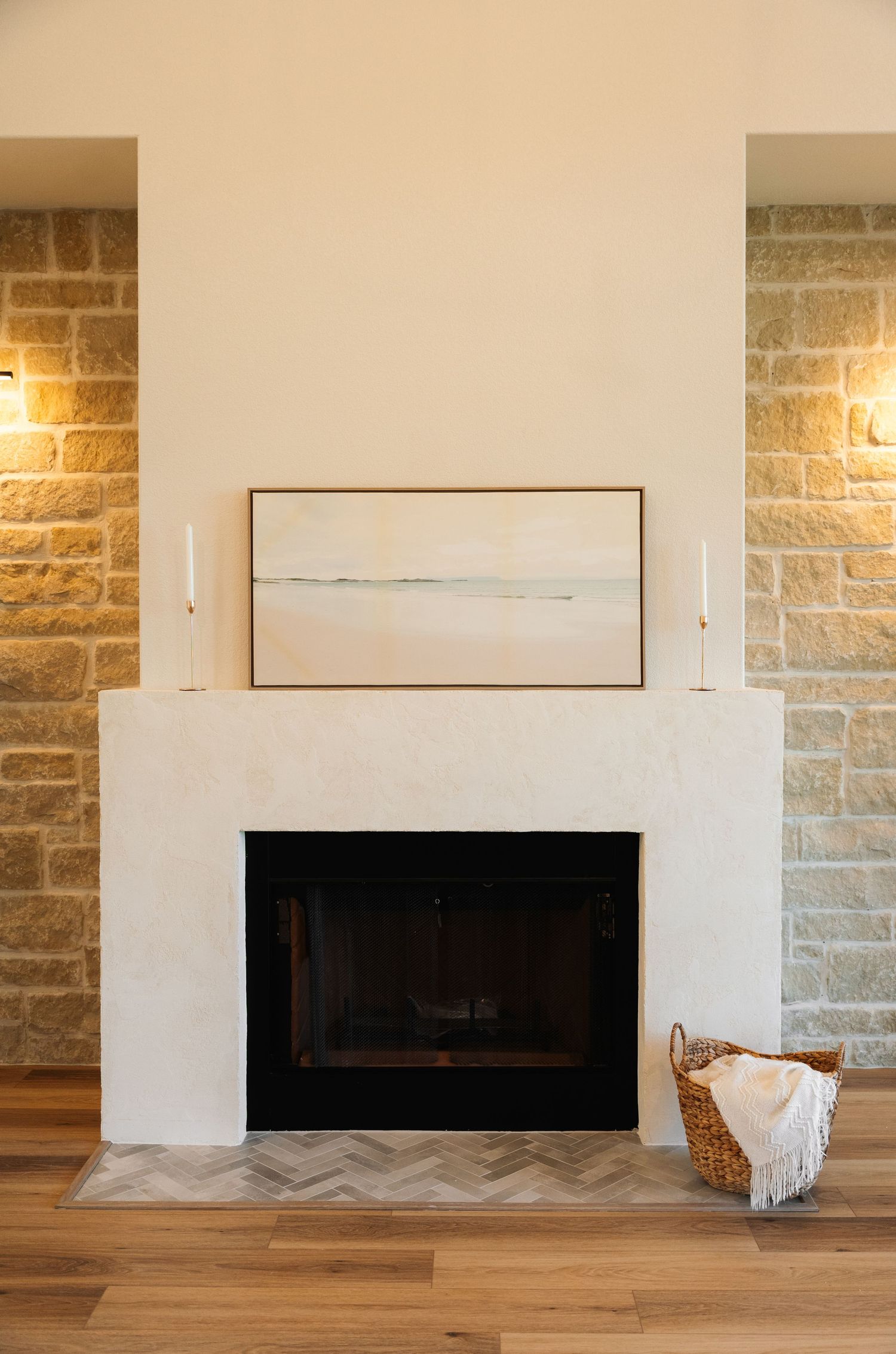 White plaster fireplace with herringbone tile hearth, abstract art, candles, and wicker basket in a stone accent wall living room.