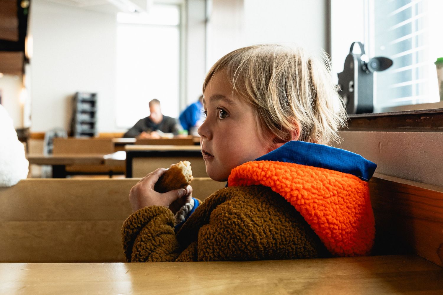 A young blonde child in a colorful fleece jacket eats a snack while sitting in a restaurant booth.