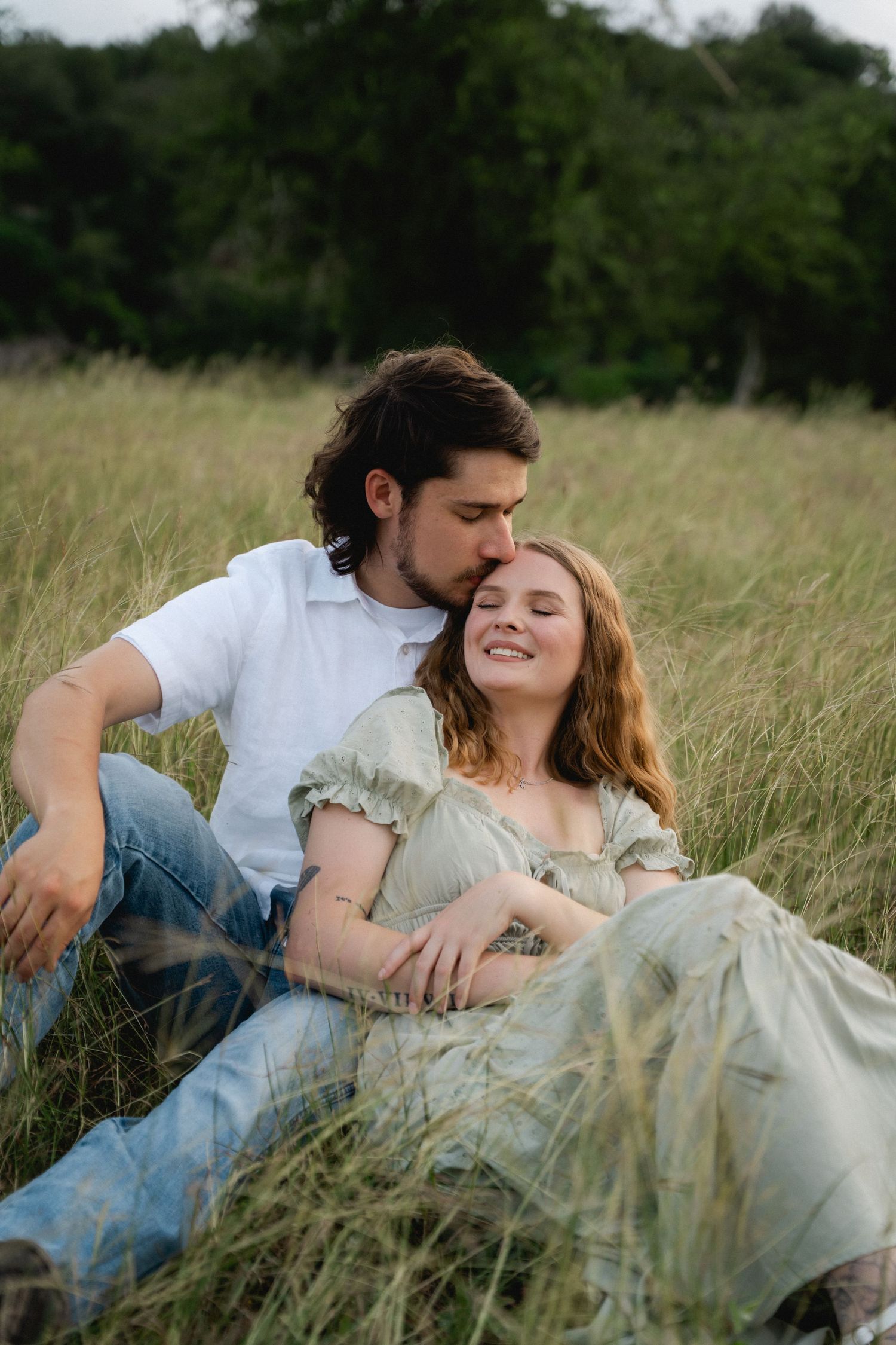 Couple sitting in tall grass field, man in white shirt kissing woman in sage green dress on her temple.
