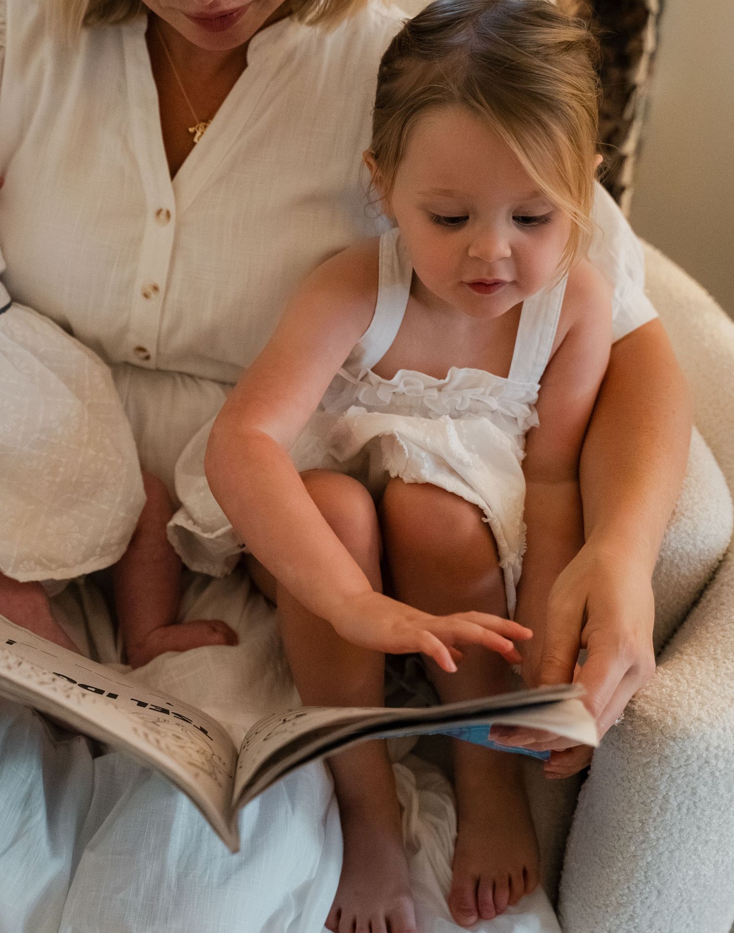 Young toddler girl sitting on mothers lap reading a picture book in a cozy white chair.