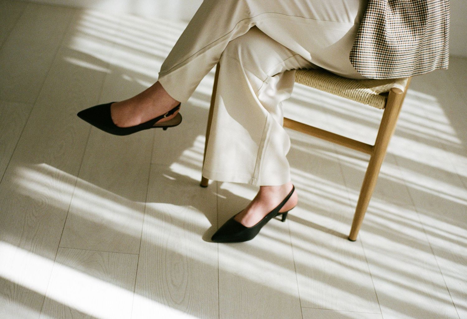 Close-up of woman's legs in beige wide-leg trousers and black kitten heel mules seated on wooden chair.